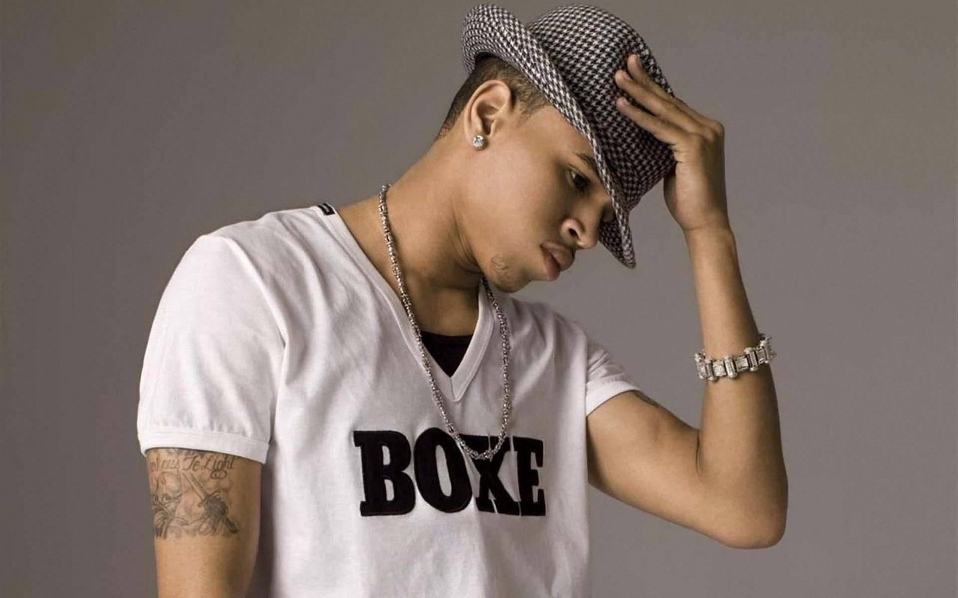 Chris Brown 桌布 壁紙 — 一名身穿白色「BOXE」T恤和格紋帽的年輕男子，在純灰色背景前低頭調整帽子。