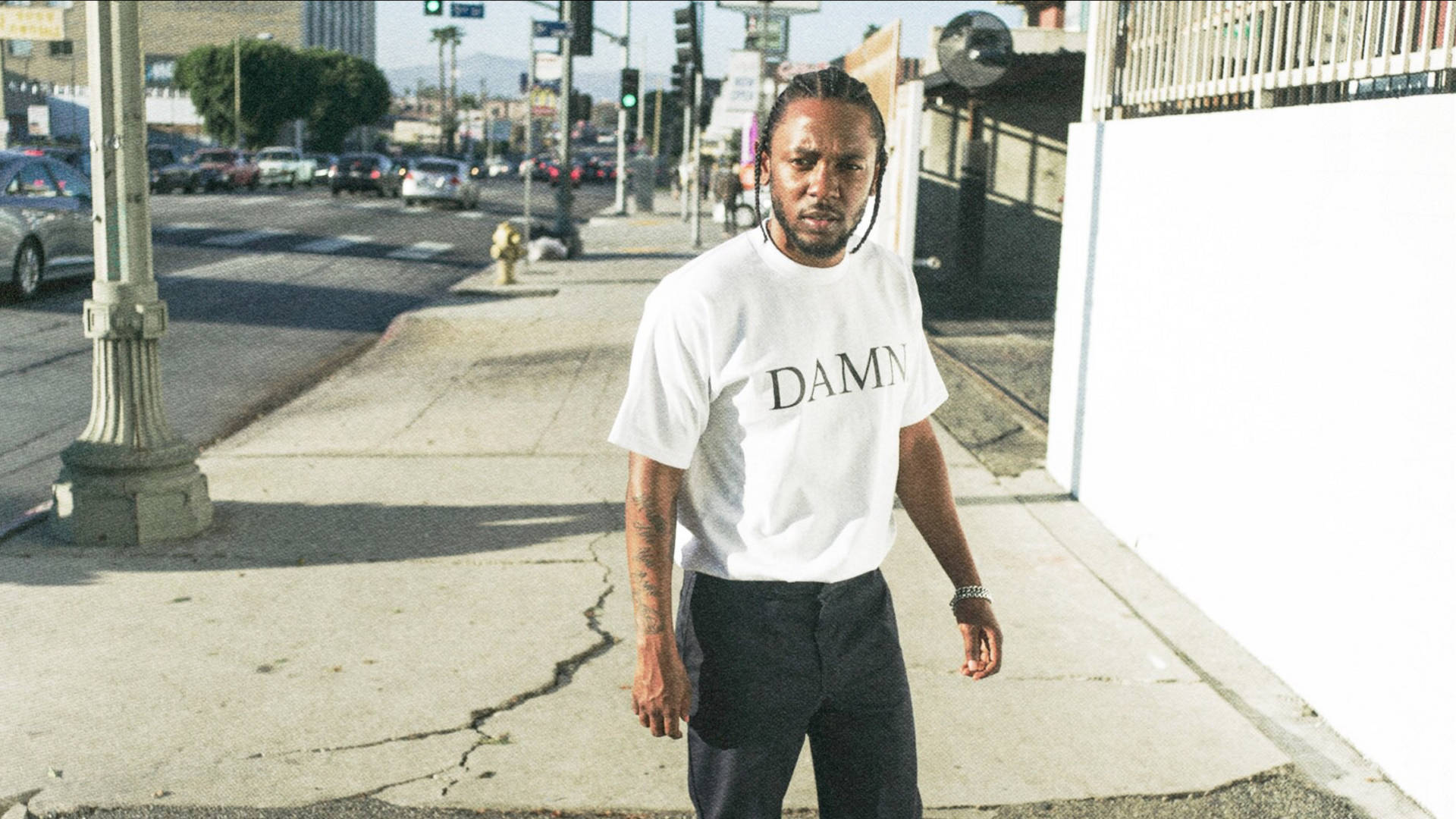 Kendrick 桌布 壁紙 — 一名身穿白色 'DAMN' T恤的男子自信地走在陽光普照的城市人行道上，背景有汽車和路燈。