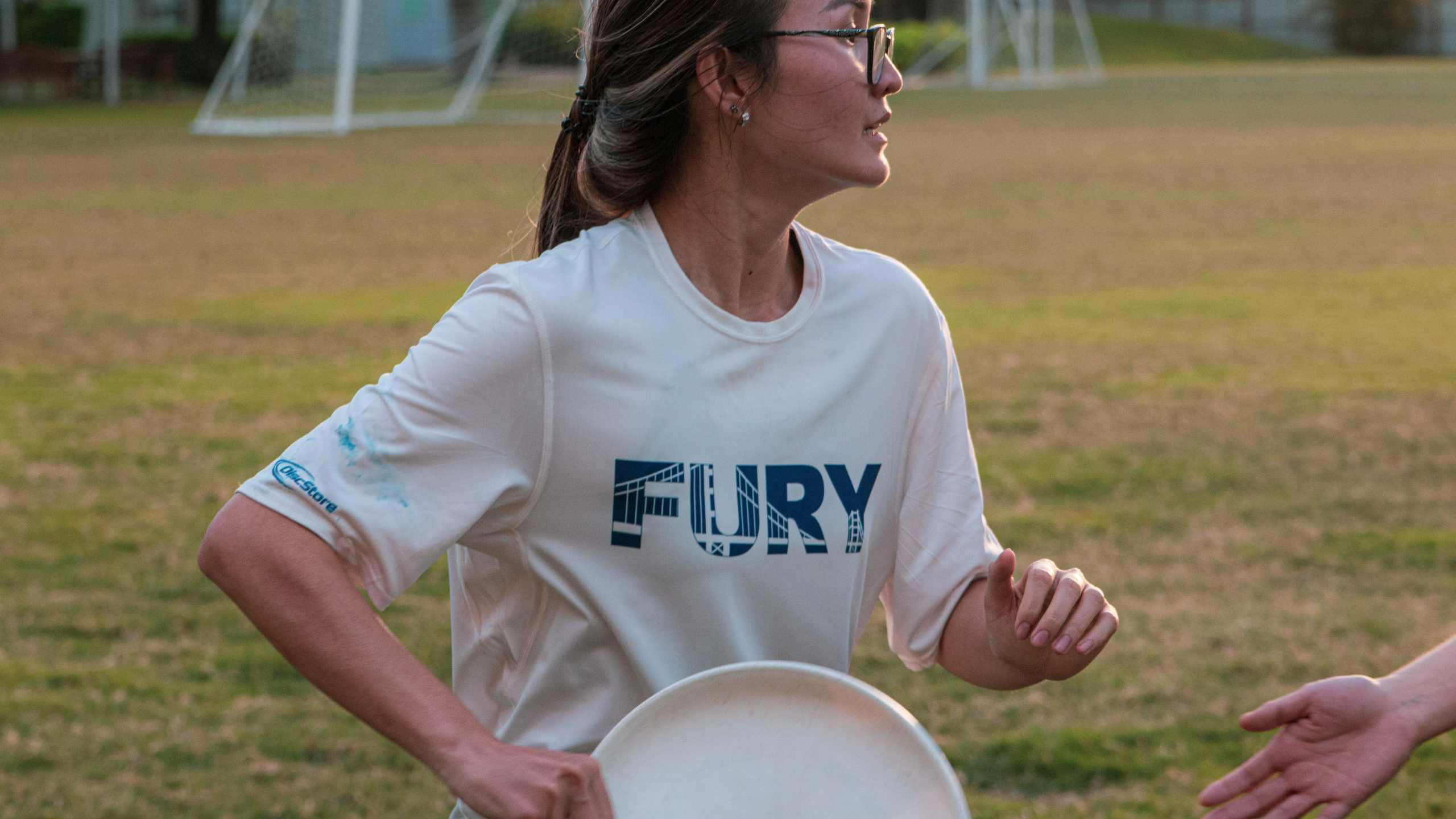 運動 桌布 壁紙 — 一名身穿白色「FURY」T恤的女性在黃金時段的草地上拿著飛盤，背景有足球球門。