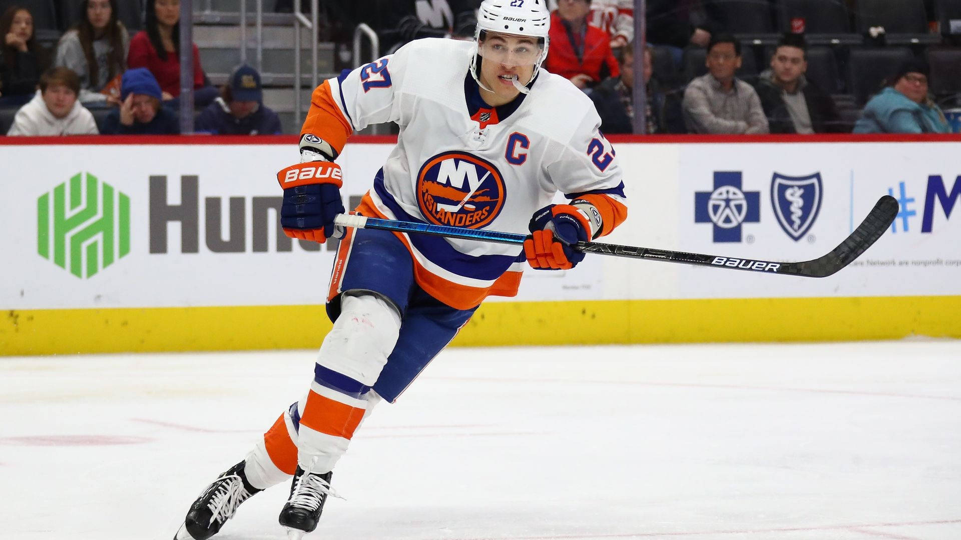 New York Islanders 桌布 壁紙 — 一名身穿白色、橙色和藍色球衣的紐約島人冰球球員在比賽期間帶著球桿在冰場上激烈滑行。