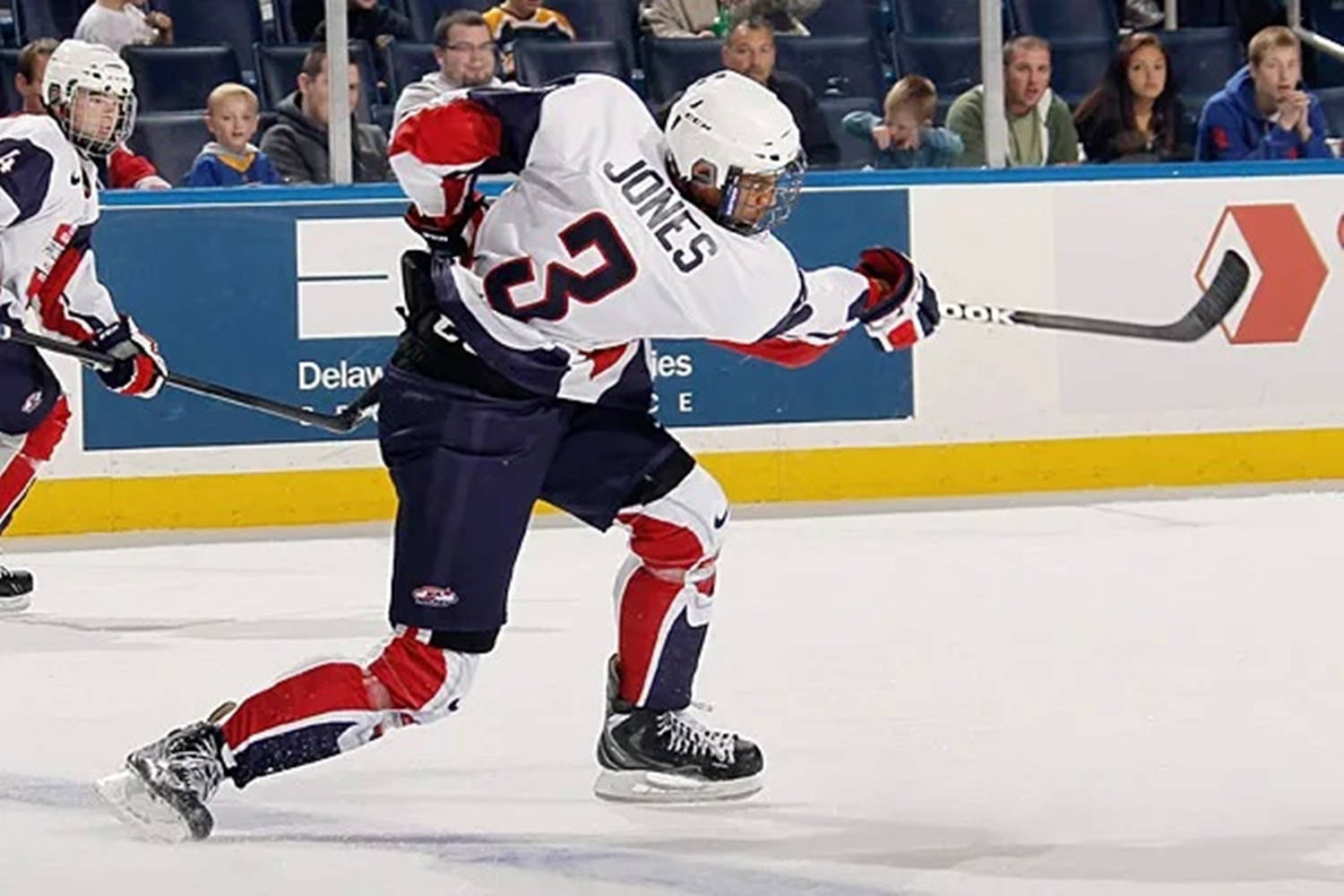 hockey 桌布 壁紙 — 一名身穿白色、紅色和藍色裝備的冰球運動員在比賽中激烈地溜冰，球桿高舉。