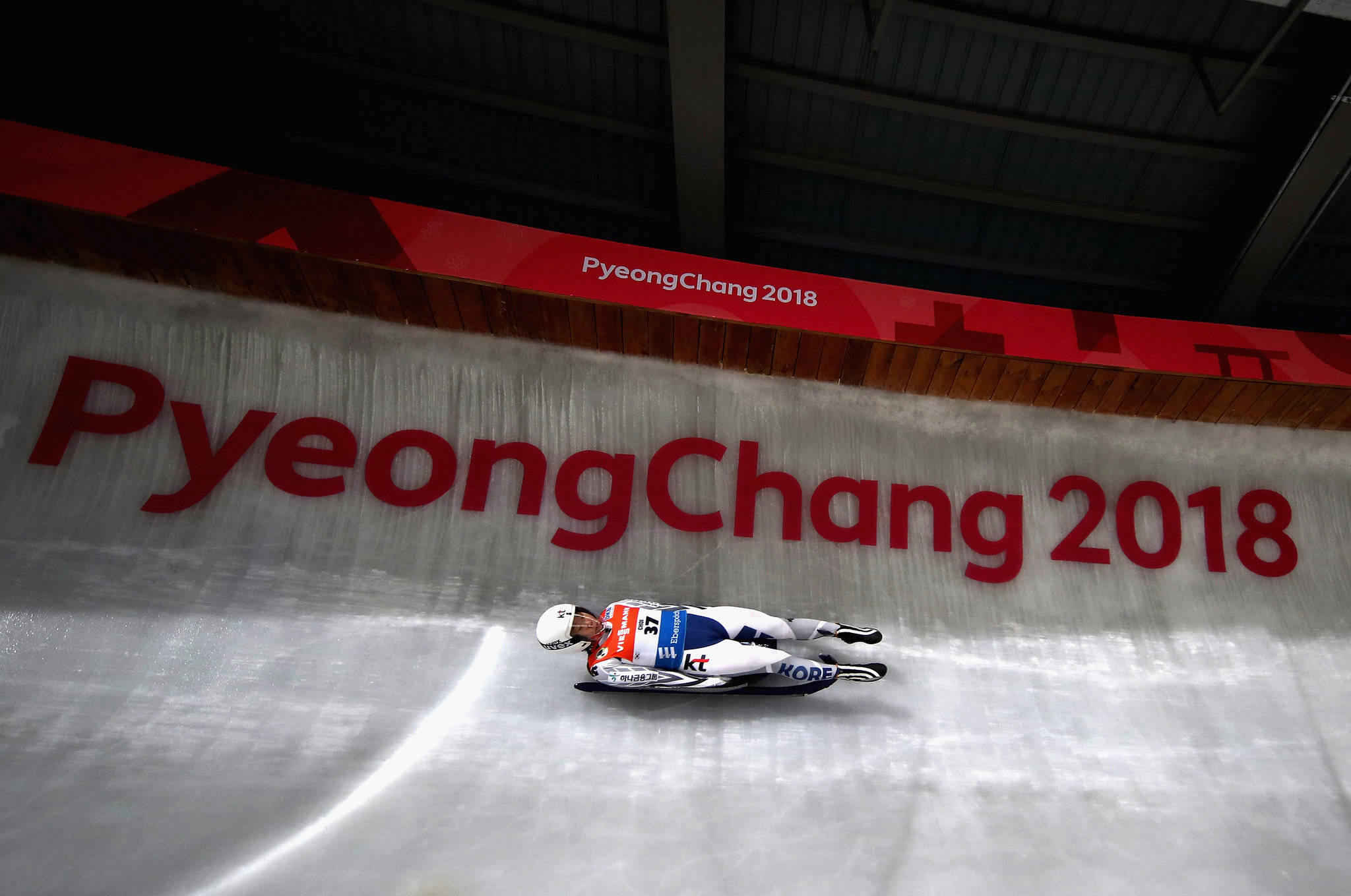 Athletic 桌布 壁紙 — 一名身穿白色、藍色和紅色套裝的雪橇運動員在冰道上飛馳，上方有「PyeongChang 2018」標誌。