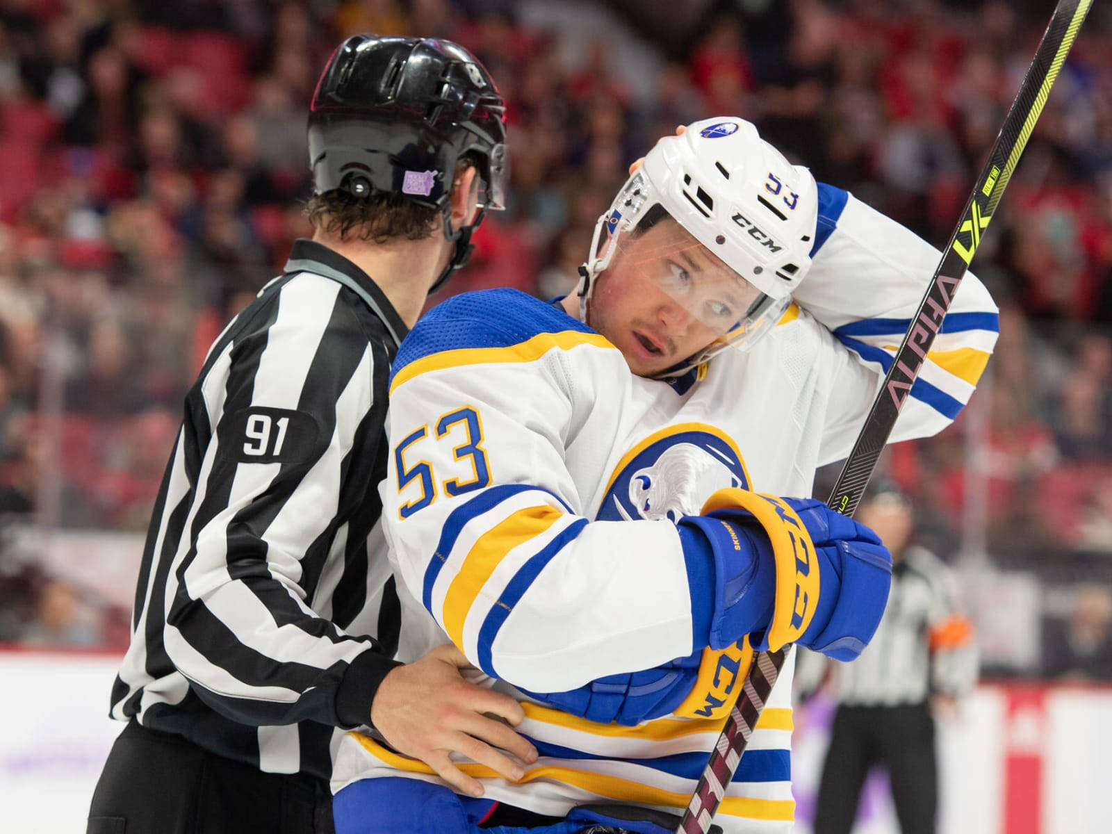 hockey 桌布 壁紙 — 一名身穿白色、藍色和黃色球衣的53號冰球運動員，在激烈的比賽中被黑白條紋的裁判攔住。