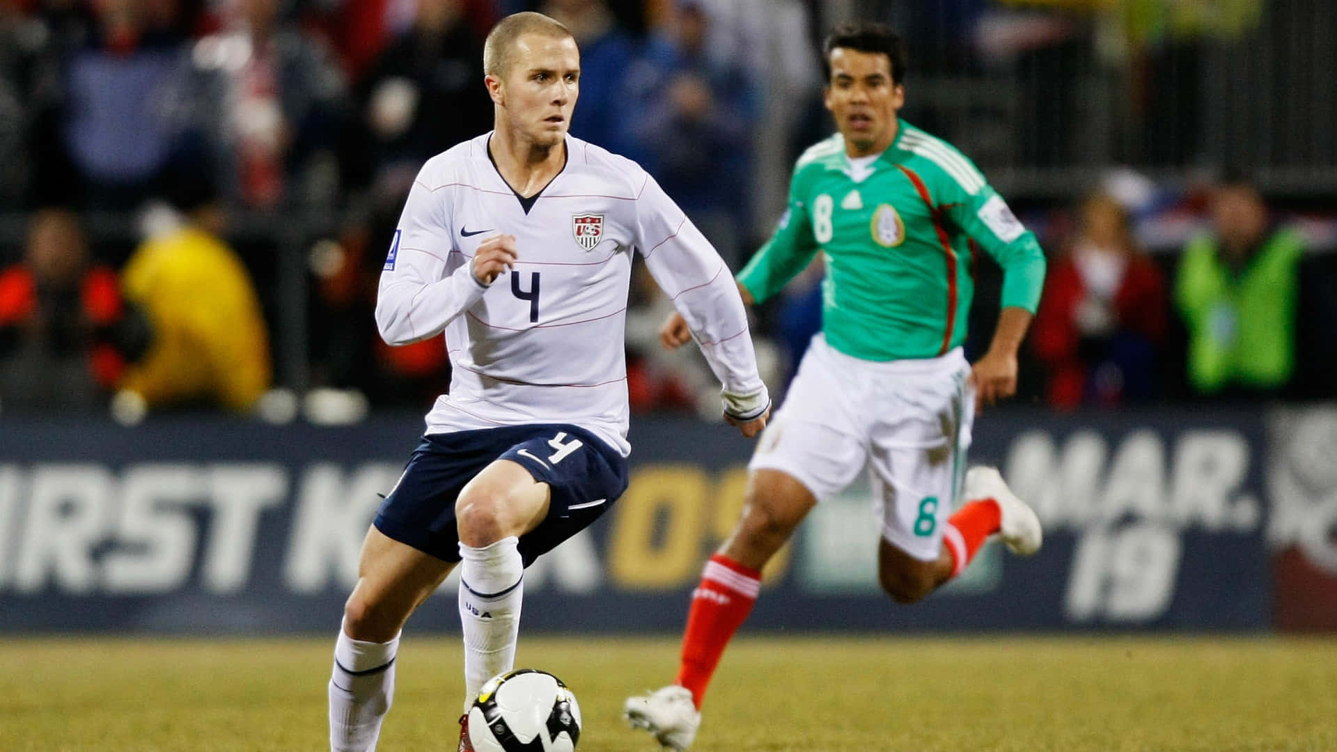 Football Players 桌布 壁紙 — 一名身穿白色4號球衣的美國足球員在球場上運球，一名身穿綠色球衣的墨西哥球員在追趕他。