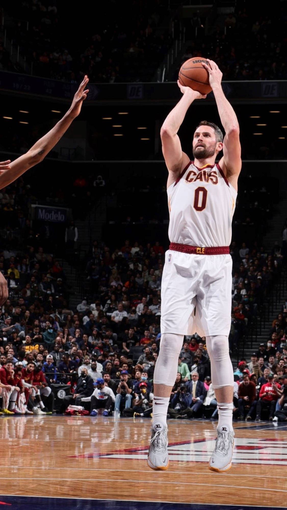 Kevin Love 桌布 壁紙 — 一名身穿白色Cavs球衣的籃球運動員跳起投籃，防守球員在擠滿觀眾的場館中伸手攔截。