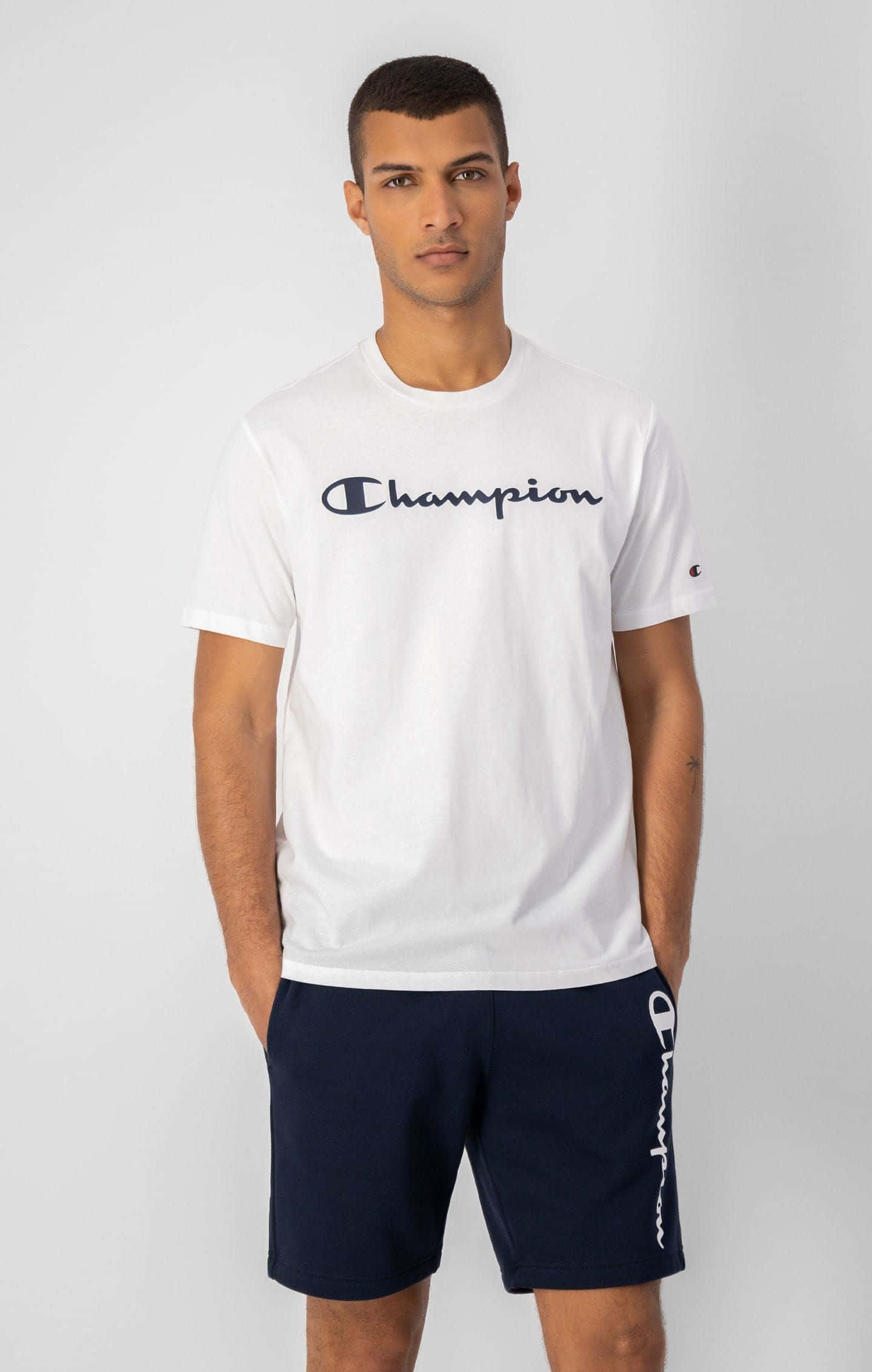 Champion 桌布 壁紙 — 一名身穿白色Champion T恤和深藍色短褲的男子自信地站在純灰色背景前。