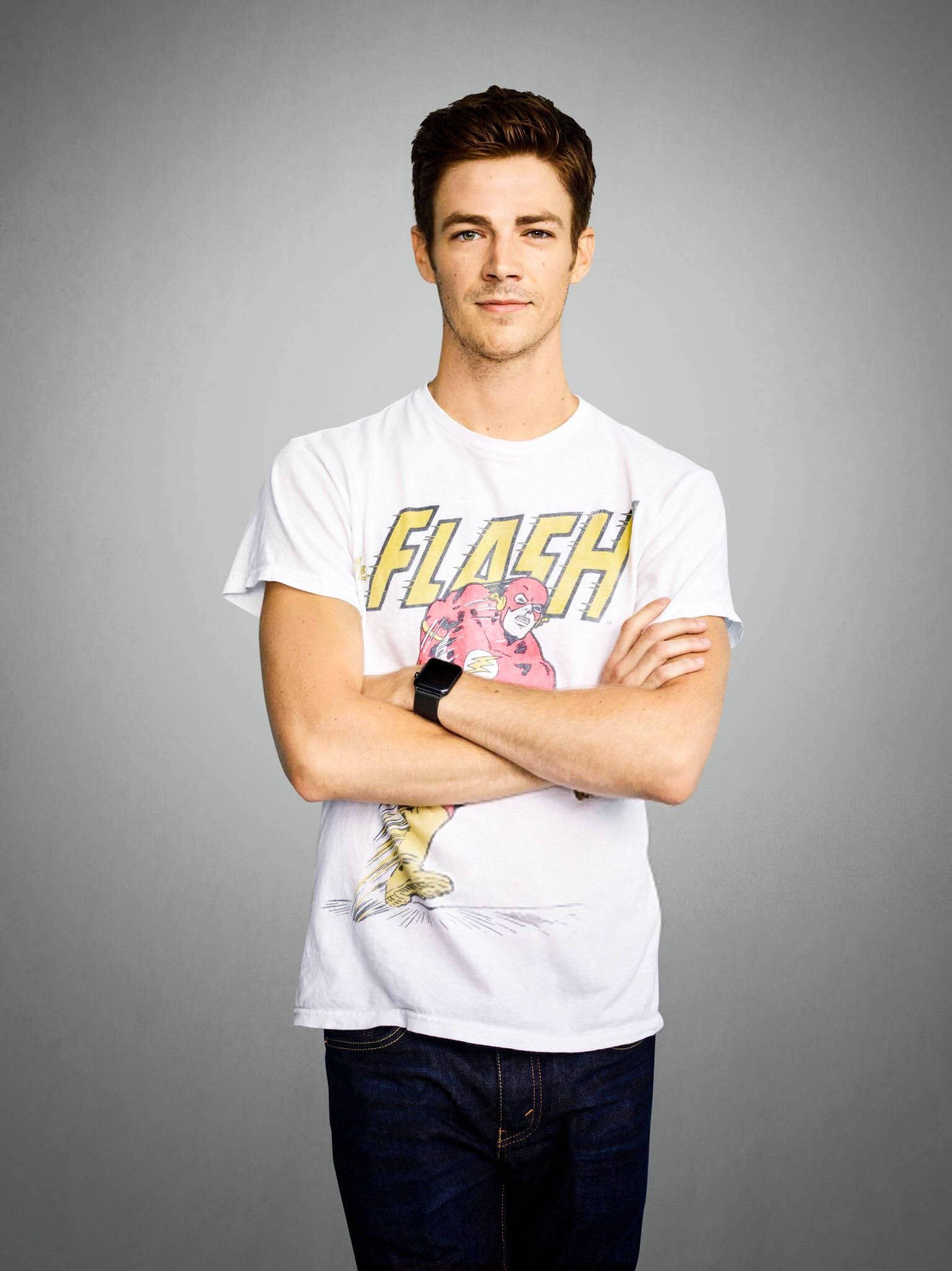 Grant Gustin 桌布 壁紙 — 一名身穿白色Flash T恤和牛仔褲的男子，在純灰色工作室背景前自信地交叉雙臂站立。