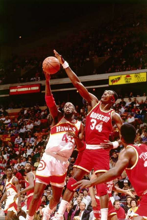 NBA 桌布 壁紙 — 一名身穿白色Hawks球衣的籃球運動員跳起投籃，而一名紅色Houston Rockets防守球員在擠滿觀眾的場館中跳起封阻。