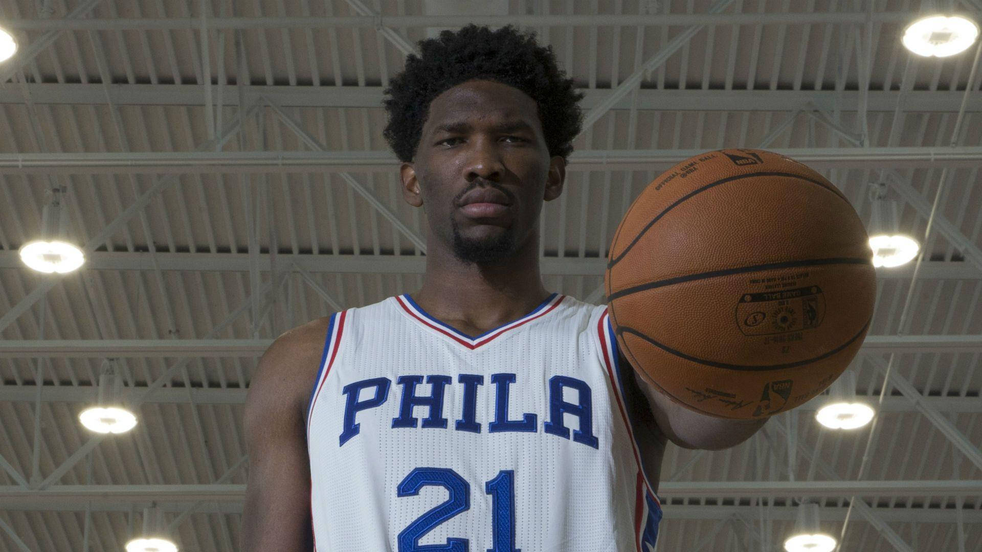 Embiid 桌布 壁紙 — 一名身穿白色PHILA球衣的籃球運動員在明亮的體育館燈光下，暴露的天花板桁架下拿著球。