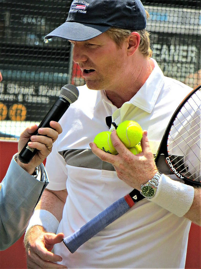 Tennis 桌布 壁紙 — 一名身穿白色Polo衫和帽子的男網球選手在球場上拿著網球和球拍，同時對著麥克風說話。