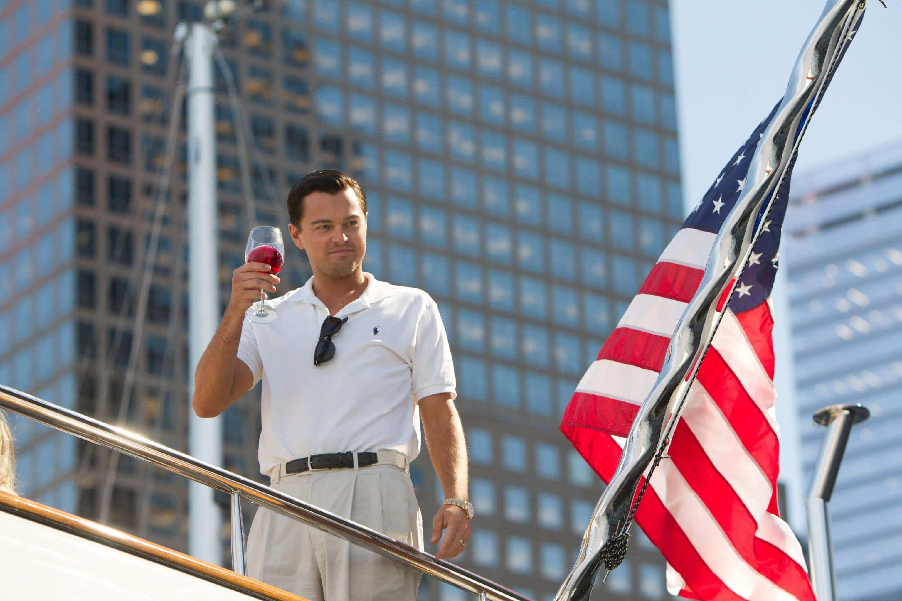 Wolf Of Wall Street 桌布 壁紙 — 一名身穿白色Polo衫的男子在遊艇甲板上品嚐紅酒，背景是美國國旗飄揚和高聳的城市摩天大樓。