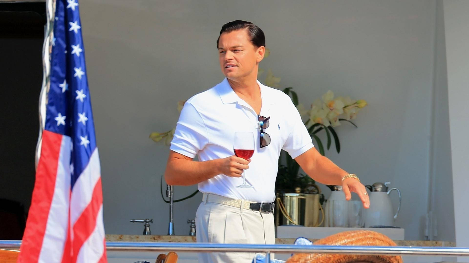 Wolf Of Wall Street 桌布 壁紙 — 一名身穿白色Polo衫的男子在陽光明媚的遊艇甲板上，旁邊有一面美國國旗，手持一杯紅酒。