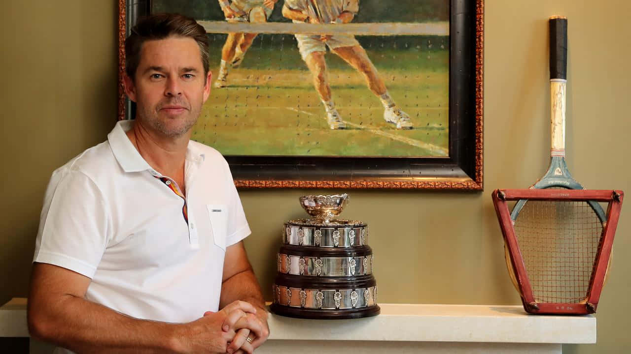 Tennis 桌布 壁紙 — 一名身穿白色Polo衫的男子站在一個大型網球獎盃和復古球拍旁邊，獎盃和球拍放在壁爐架上，身後有一幅網球畫。