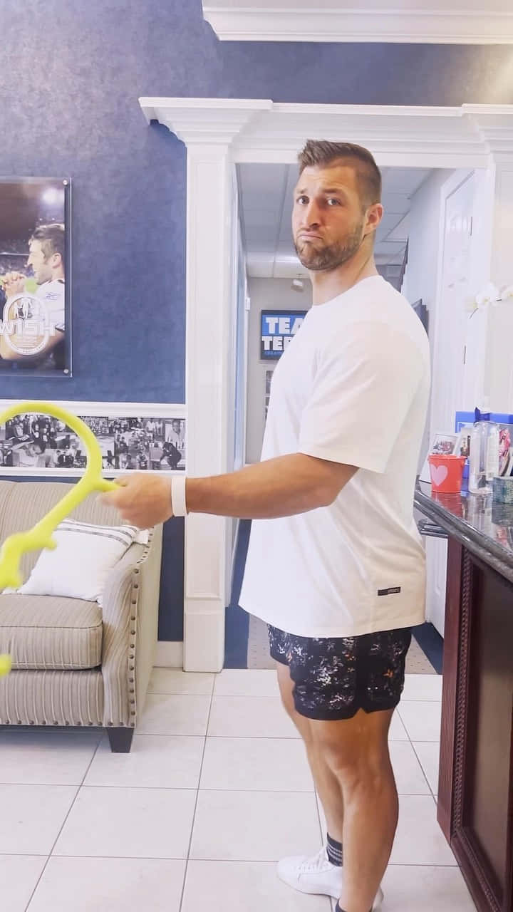 Tim Tebow 桌布 壁紙 — 一名身穿白色T恤和圖案短褲的男子，在一個鋪有瓷磚、藍色牆壁和掛有運動照片的房間裡，拿著一支亮黃色的網球拍。