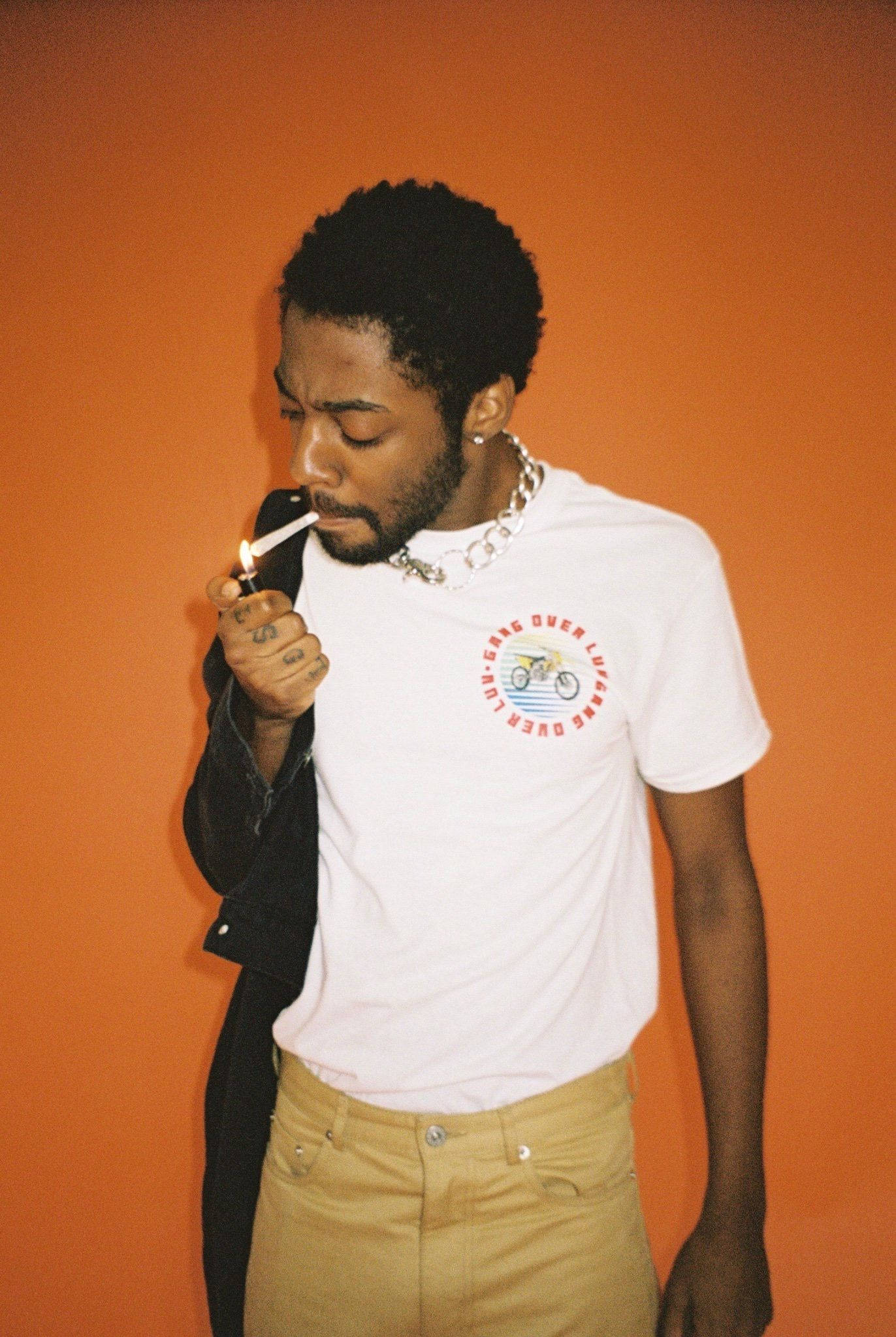 Brent Faiyaz 桌布 壁紙 — 一名身穿白色T恤和米色褲子的男子，在純橙色牆壁前點燃一支香煙。