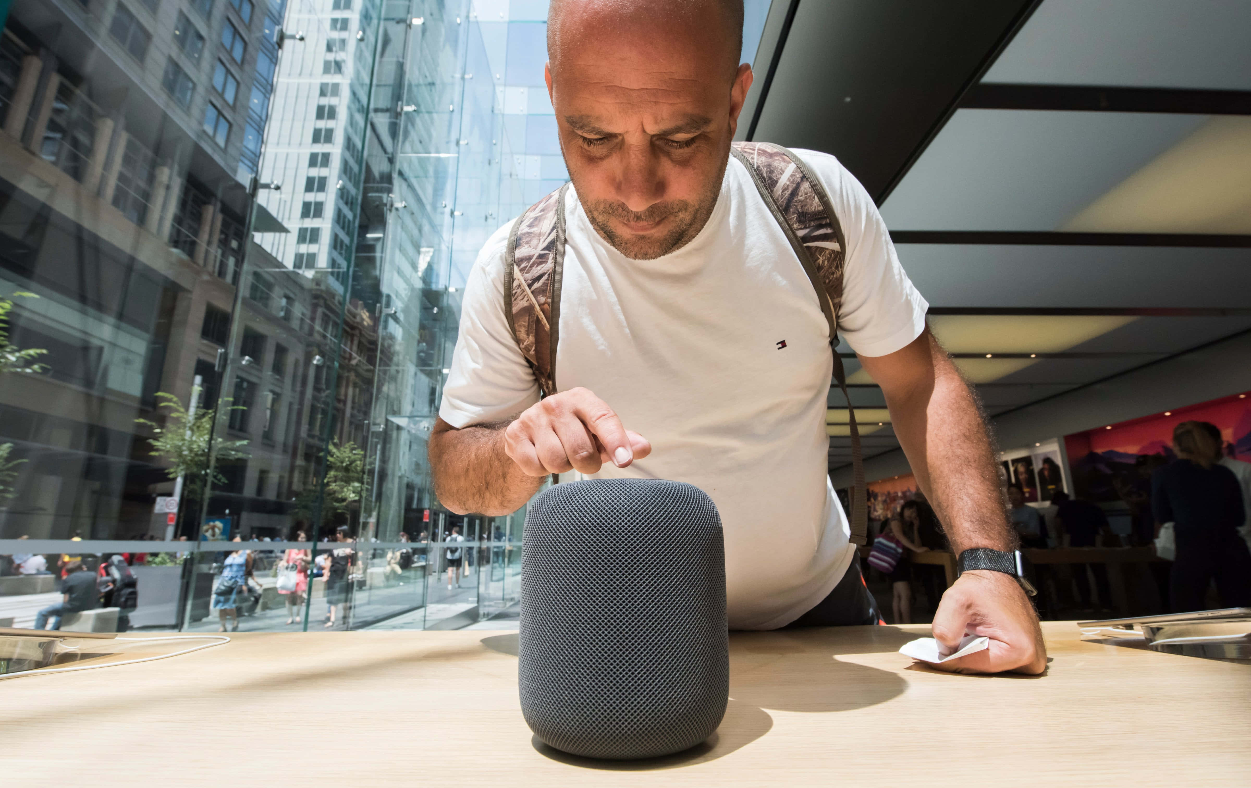 Homepod 桌布 壁紙 — 一名身穿白色T恤的男子俯身靠在木製櫃檯上，觸摸一個灰色智慧型揚聲器，店舖為現代玻璃幕牆設計。