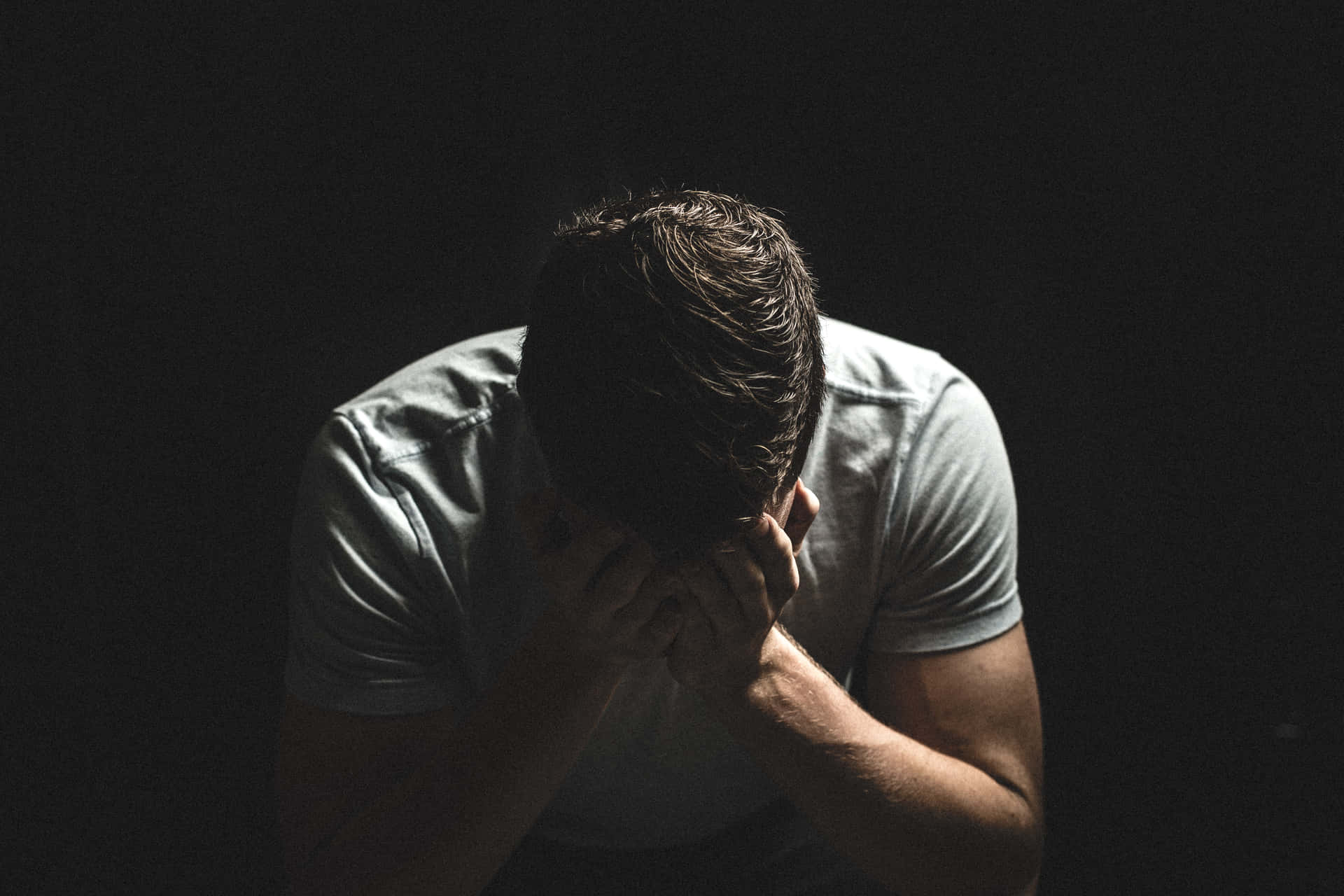Sad Face 桌布 壁紙 — 一名身穿白色T恤的男子在純黑背景下彎腰低頭，雙手抱頭。