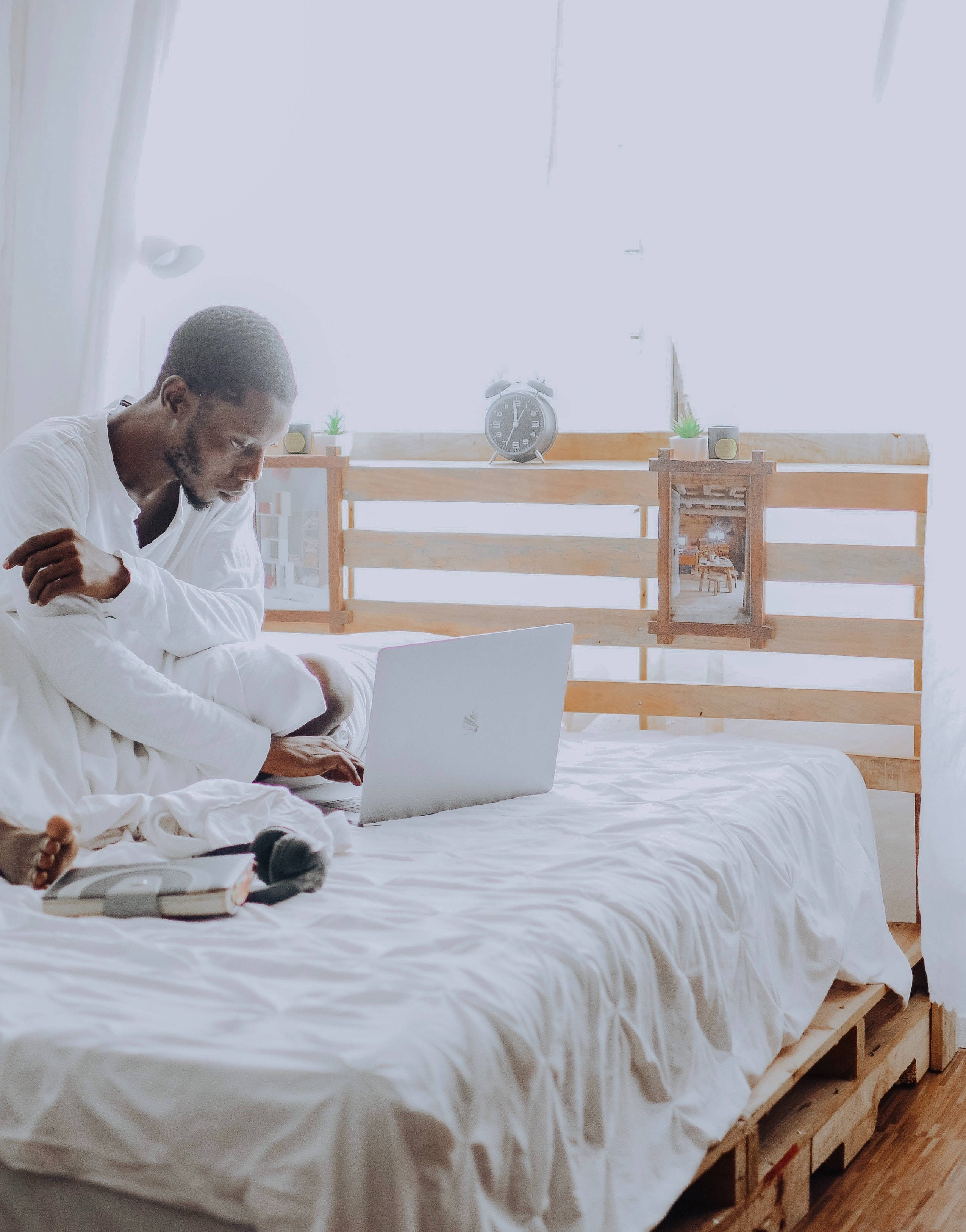 aesthetic tumblr laptop 桌布 壁紙 — 一名身穿白色休閒服的男子盤腿坐在木棧板床上，在明亮的極簡主義臥室中使用銀色筆記型電腦。