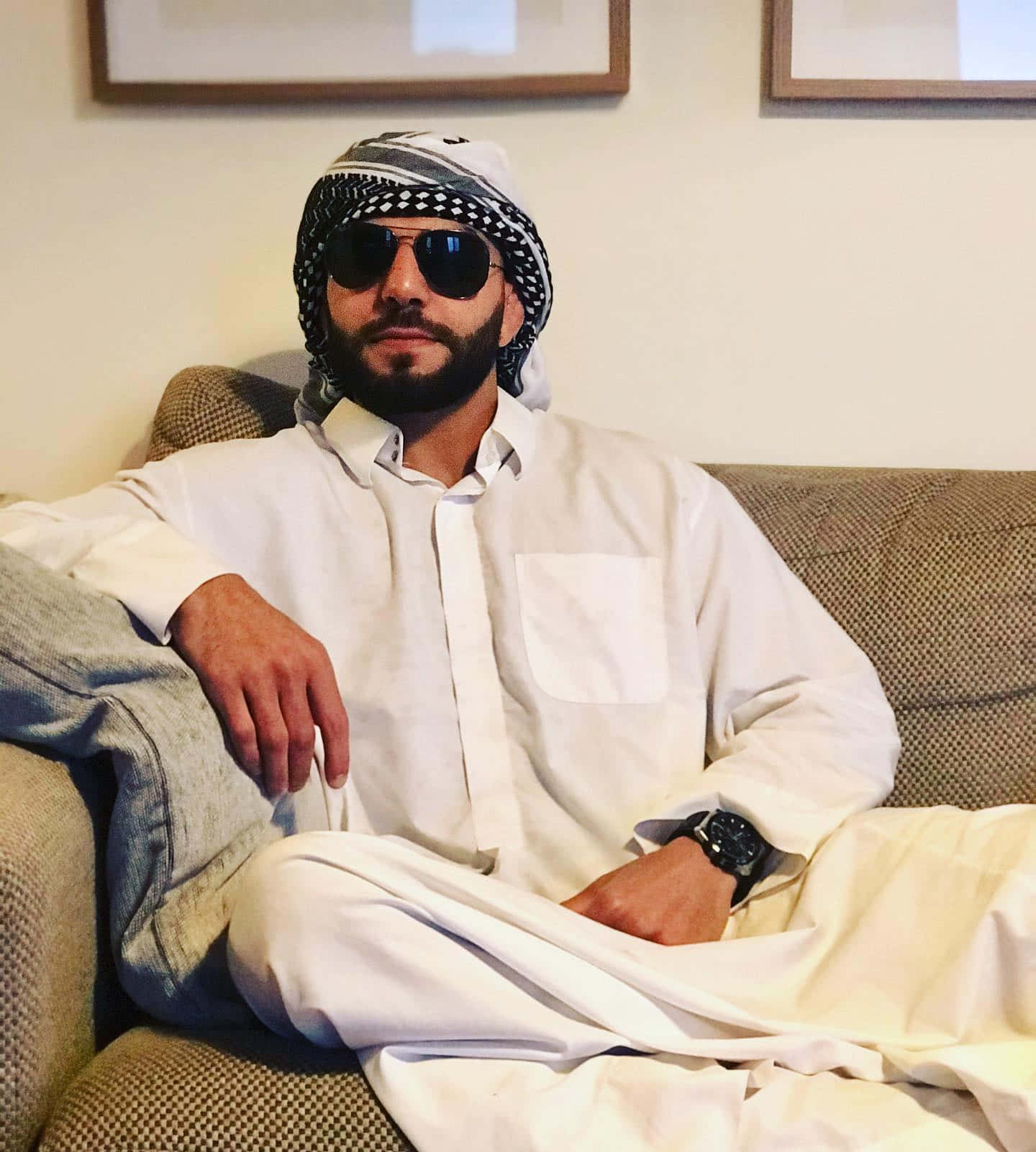 Sunglasses 桌布 壁紙 — 一名身穿白色傳統服飾、戴著太陽眼鏡的男子，自信地坐在室內一張有紋理的沙發上。