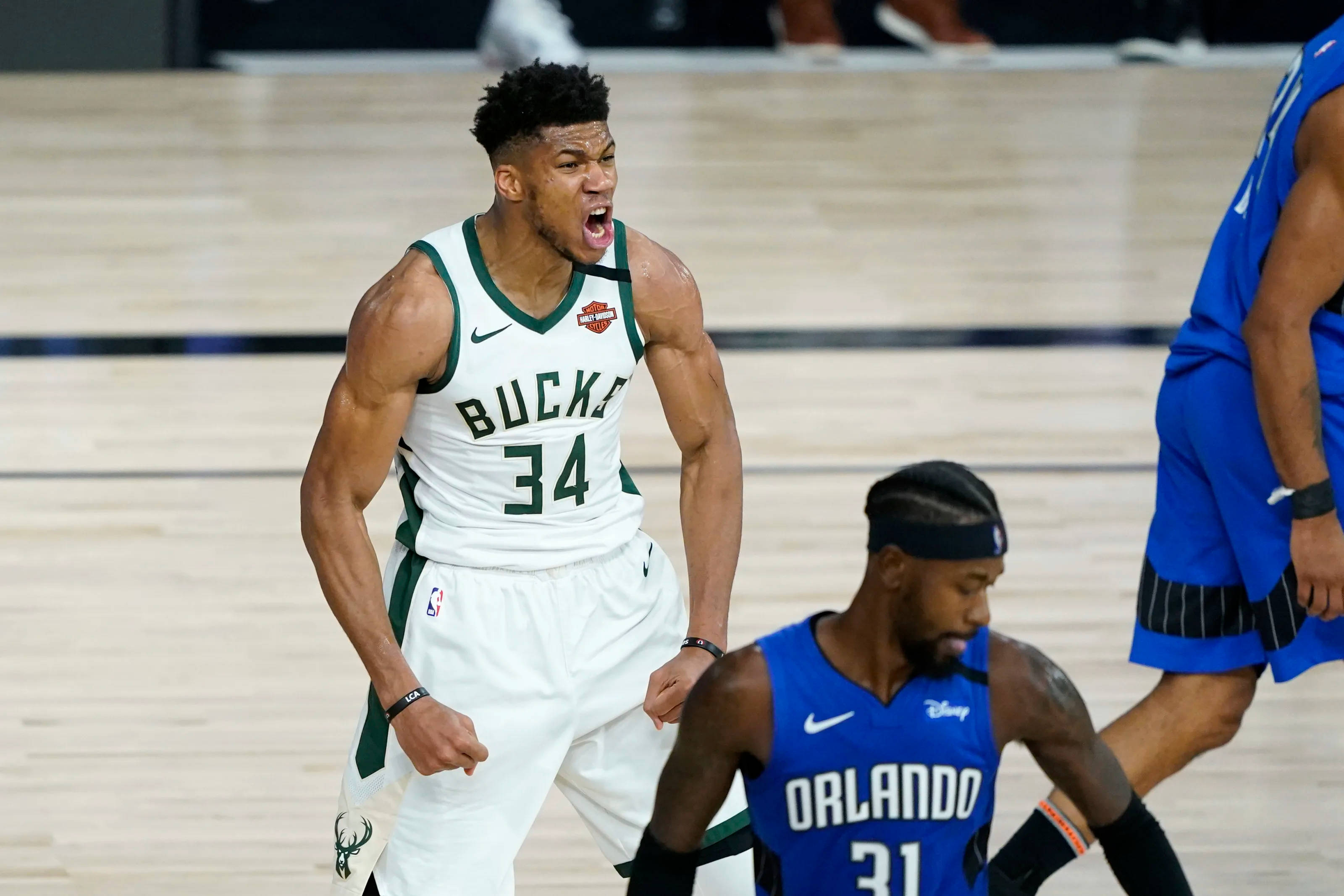 Giannis 桌布 壁紙 — 一名身穿白色公鹿球衣的籃球運動員在比賽中激烈咆哮，而身穿藍色球衣的對手則在旁觀看。