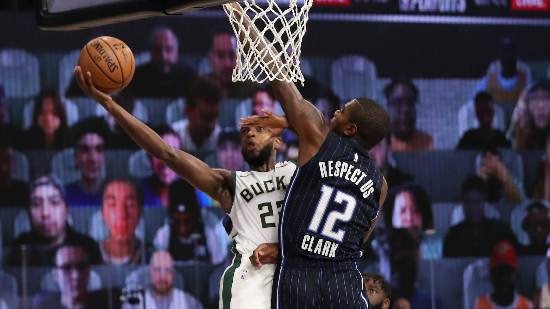 Khris Middleton 桌布 壁紙 — 一名身穿白色公鹿球衣的籃球運動員跳起投籃，而一名身穿海軍藍細條紋球衣、印有「RESPECT US」和12號的防守球員在明亮的場館燈光下封阻投籃。