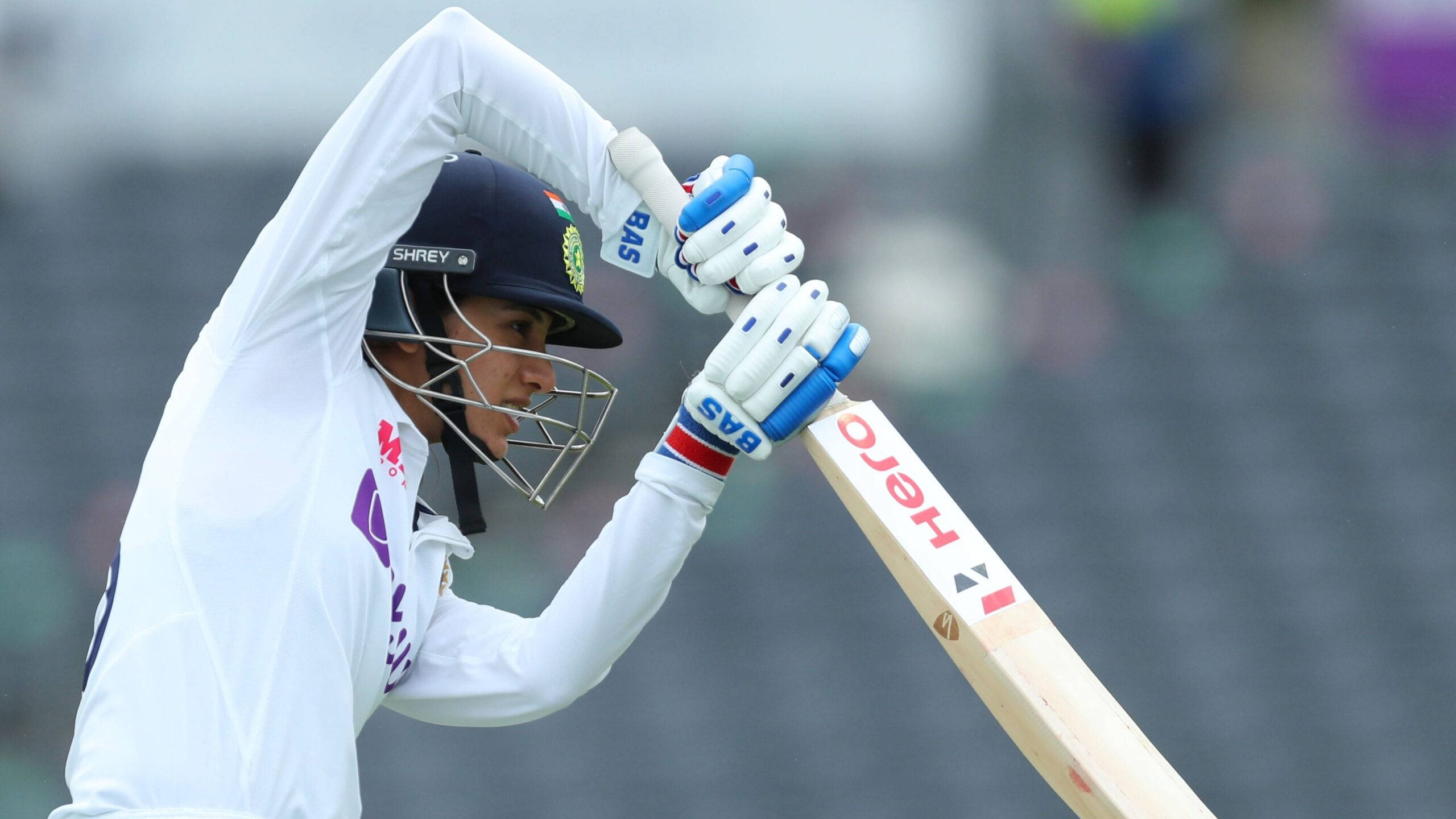 Smriti Mandhana 桌布 壁紙 — 一名身穿白色制服和頭盔的板球運動員在綠色場地上以勝利姿態舉起球棒。