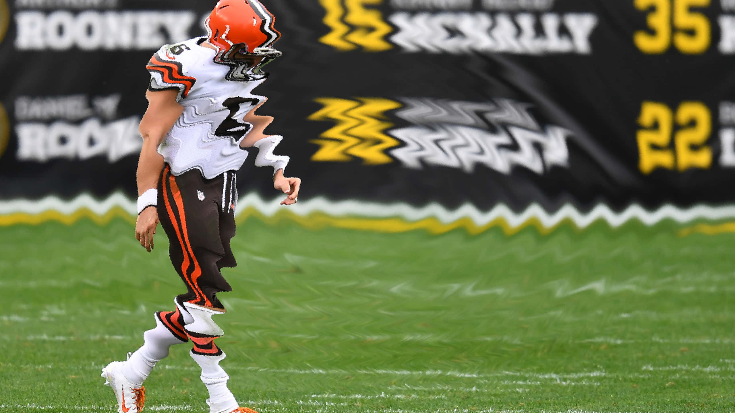 Baker Mayfield 桌布 壁紙 — 一名身穿白色和橙色球衣的克里夫蘭布朗隊美式足球員垂頭喪氣地走在綠色草地上，背景有記分板。
