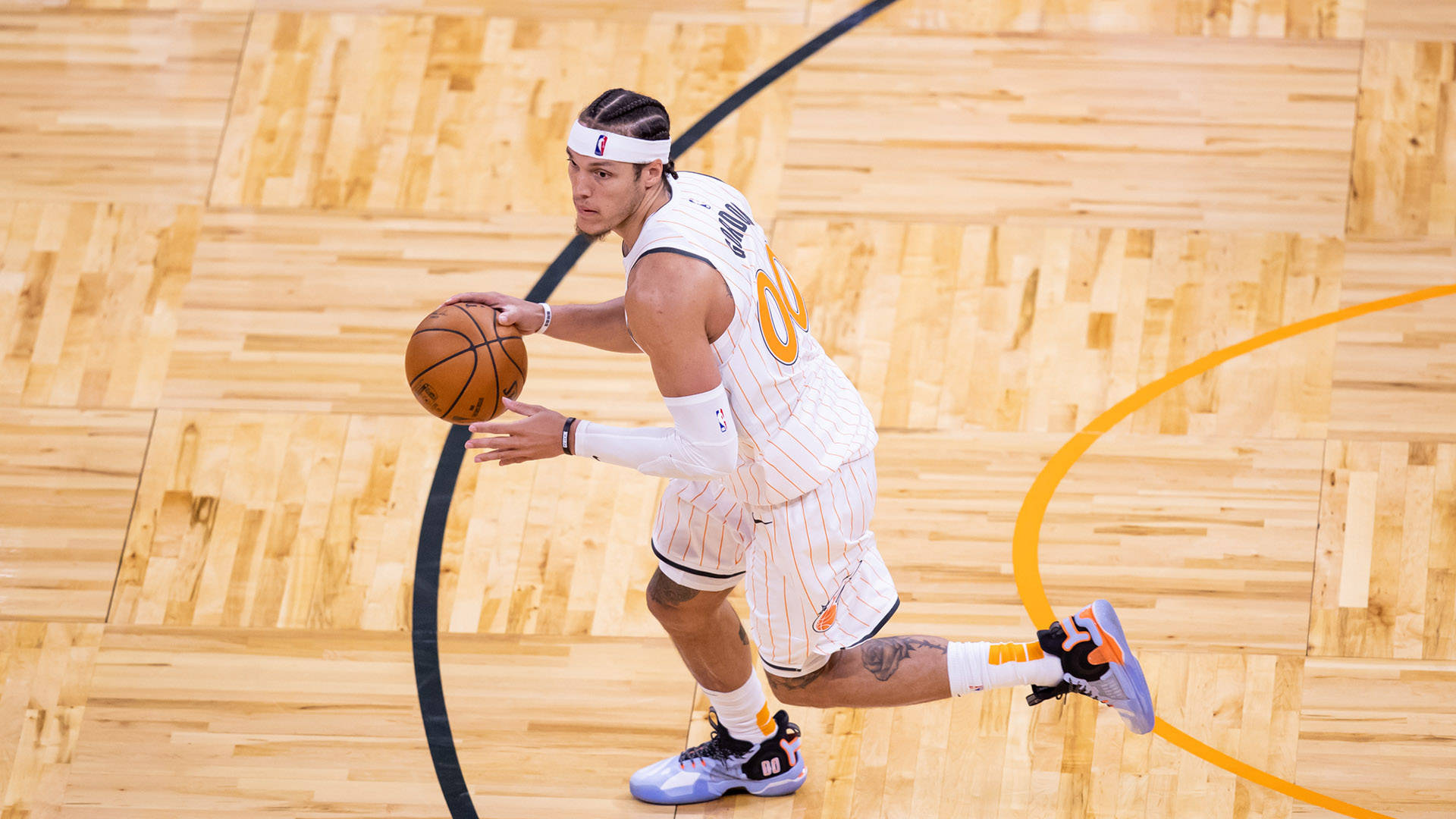 aaron gordon 桌布 壁紙 — 一名身穿白色和橙色球衣的籃球運動員在明亮的場館燈光下，在拋光木地板上低運球。