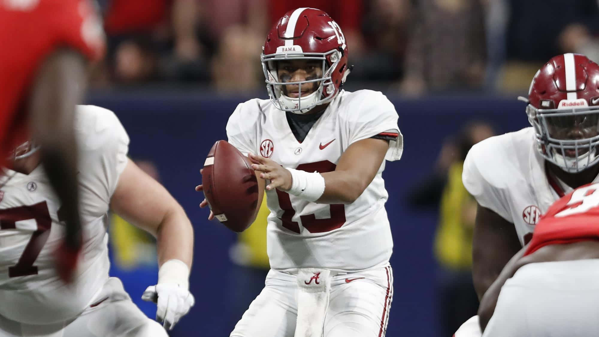 Tua Tagovailoa 桌布 壁紙 — 一名身穿白色和深紅色裝備的美式足球四分衛在體育場燈光下的夜間比賽中準備投球。