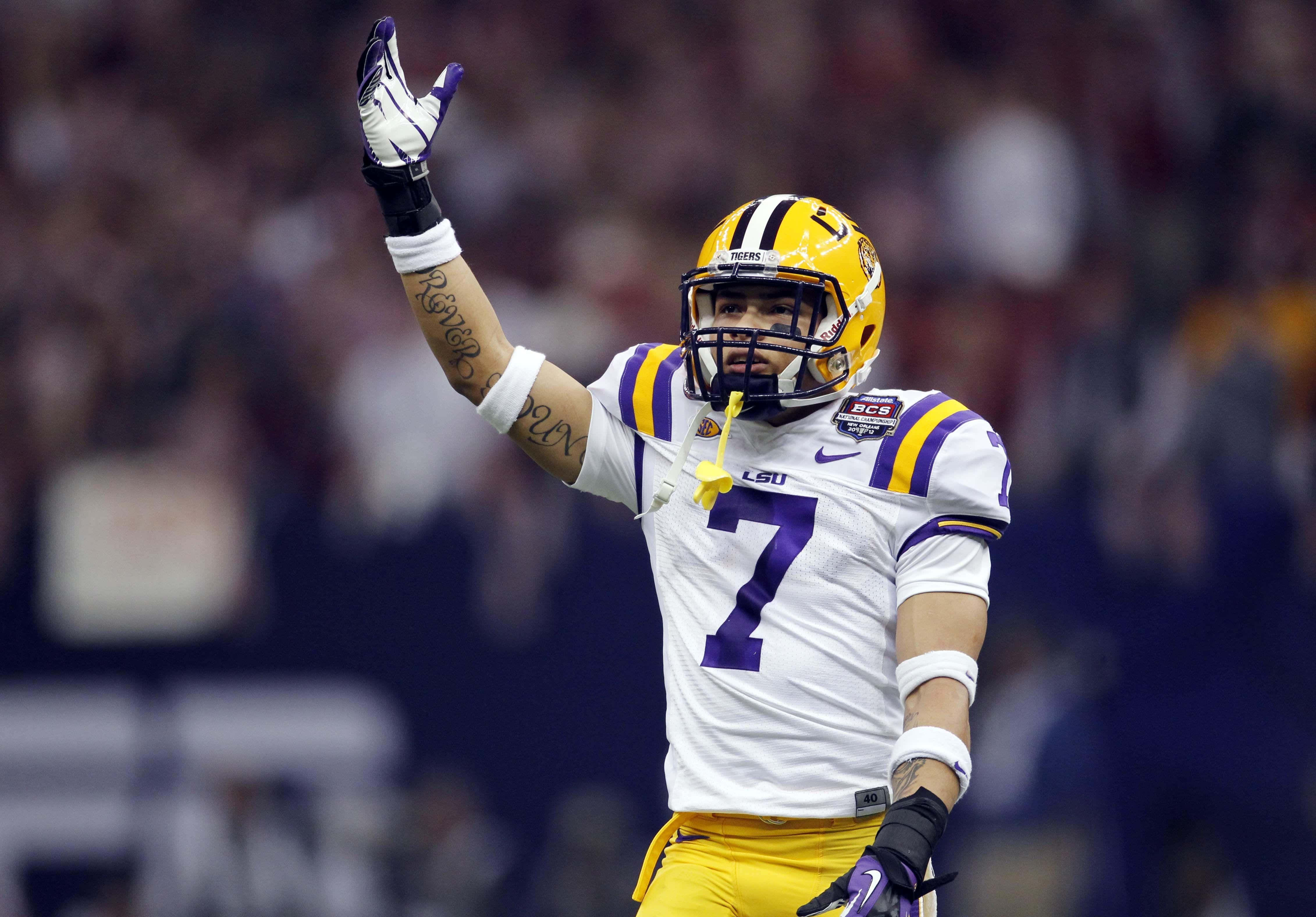 Tyrann Mathieu 桌布 壁紙 — 一名身穿白色和紫色LSU球衣的足球員在體育場燈光下的比賽中舉起戴手套的手慶祝。
