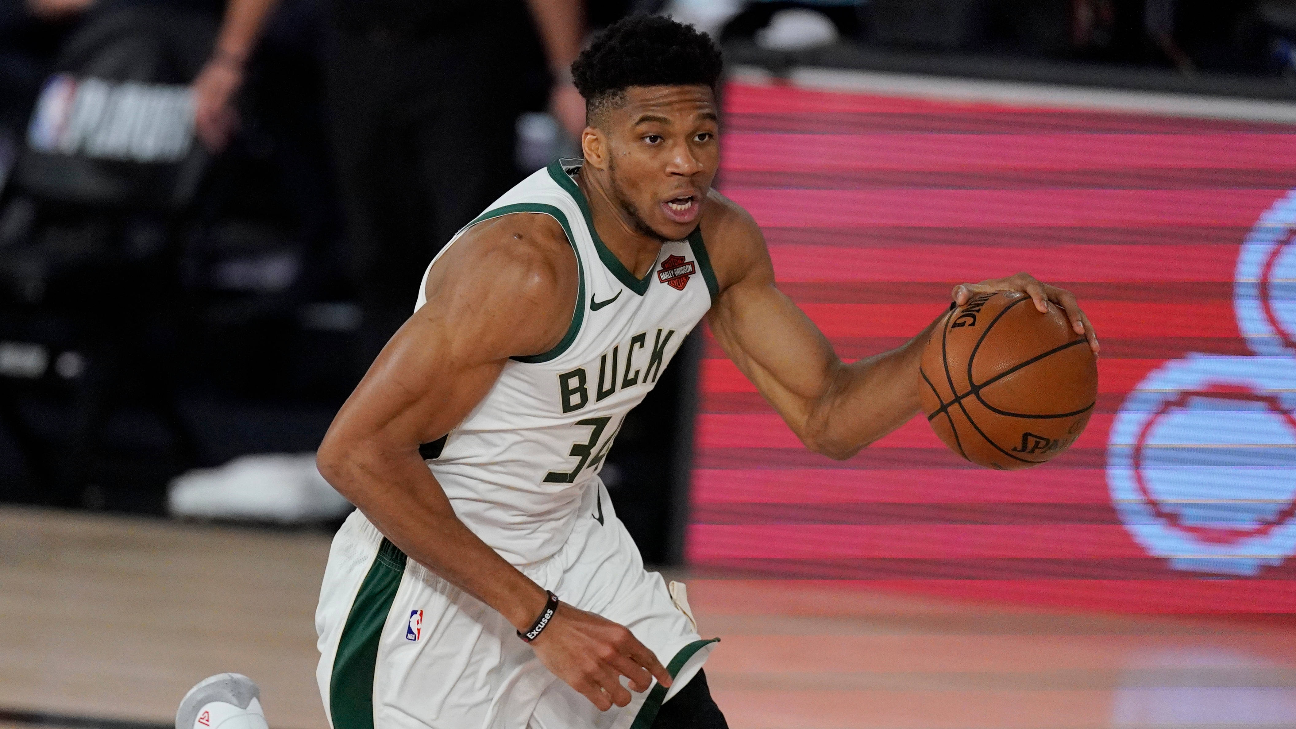 Giannis 桌布 壁紙 — 一名身穿白色和綠色公鹿球衣的籃球運動員在明亮的場館燈光下，在比賽中激烈運球。