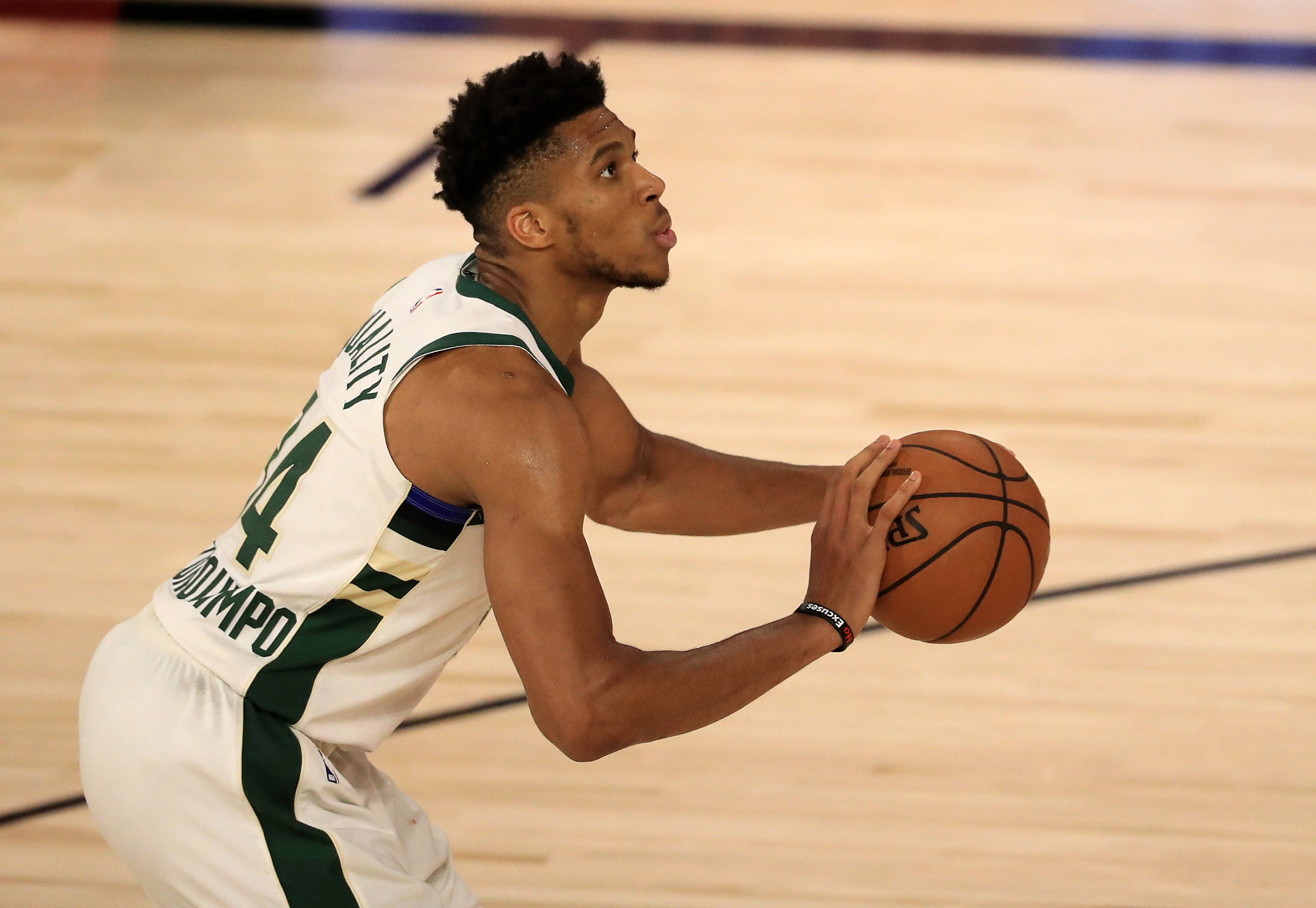 Giannis 桌布 壁紙 — 一名身穿白色和綠色球衣的籃球運動員在明亮的場館燈光下，專注地在木地板上運球。