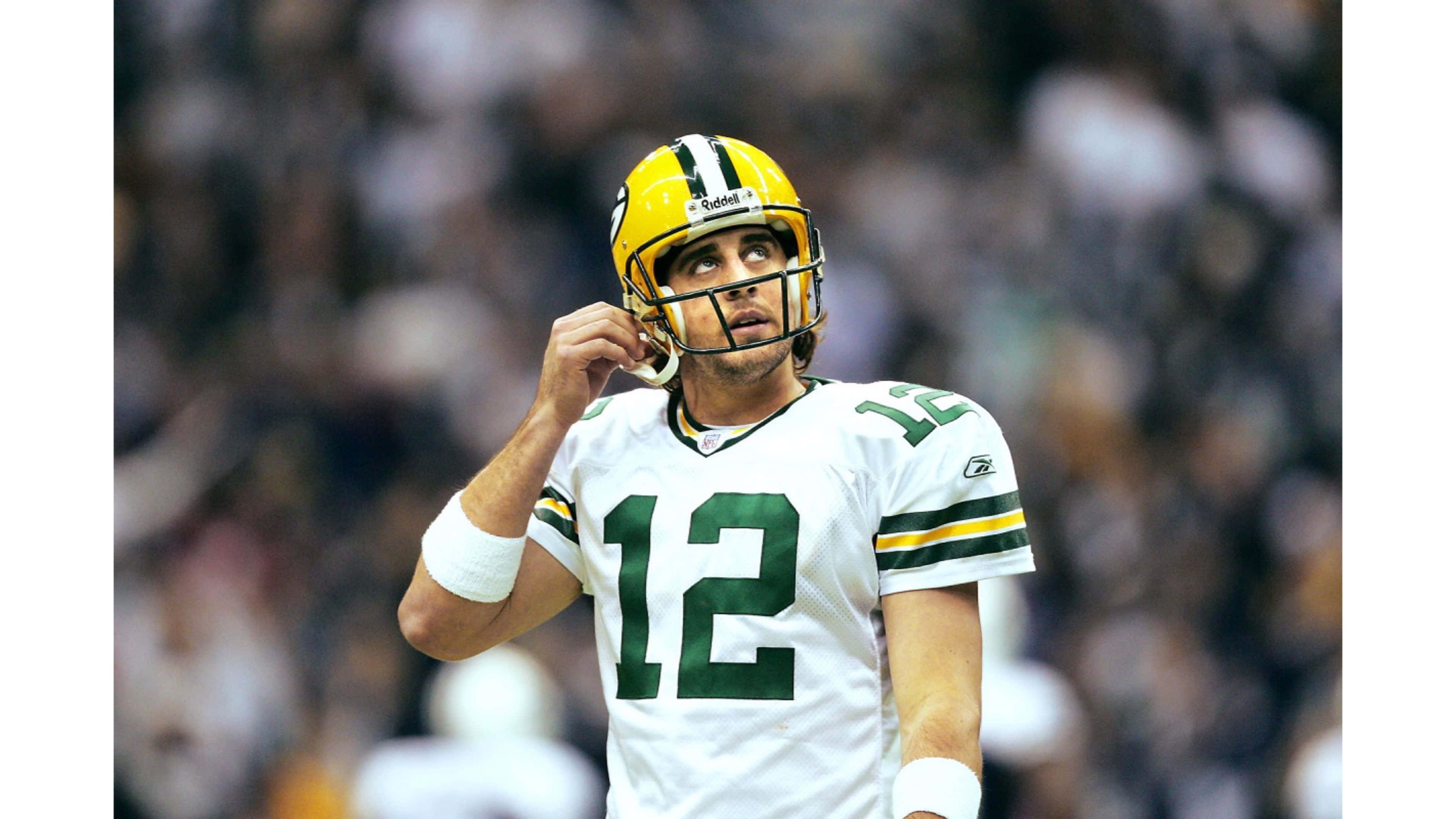 Aaron Rodgers 桌布 壁紙 — 一名身穿白色和綠色球衣的綠灣包裝工四分衛在比賽中調整他的黃色頭盔，背景是模糊的體育場人群。