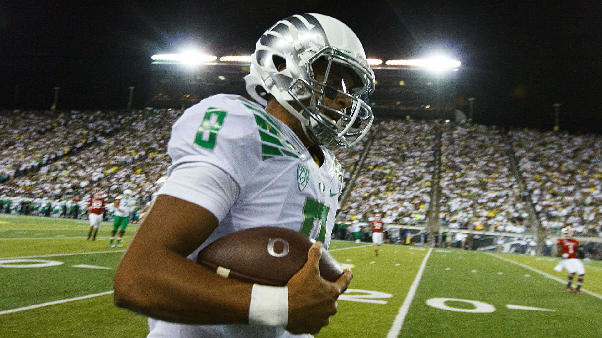 Oregon Ducks 桌布 壁紙 — 一名身穿白色和綠色球衣的足球員在球場燈光下抱著球，身後是擠滿的人群。