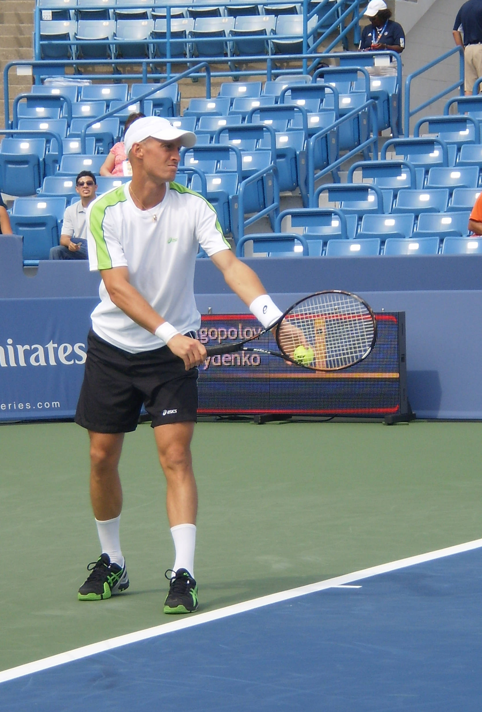 Tennis 桌布 壁紙 — 一名身穿白色和綠色裝備的男網球選手準備在藍綠色球場上擊打黃色球，身後是空的藍色座位。
