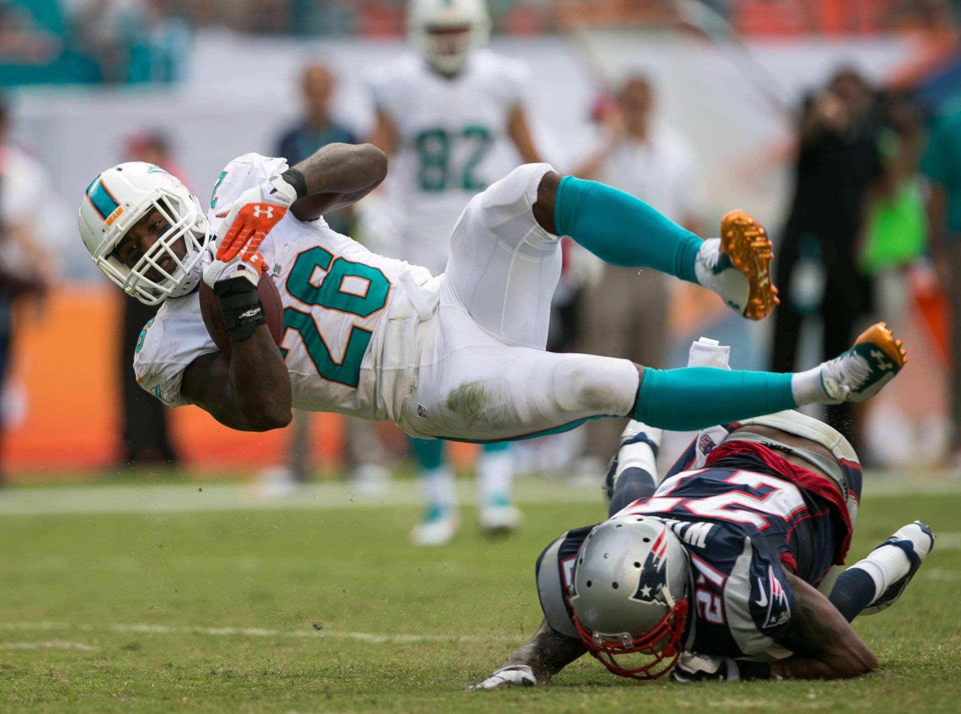 dolphins 桌布 壁紙 — 一名身穿白色和藍綠色球衣的邁阿密海豚隊球員向前飛撲，緊緊抓住橄欖球，而新英格蘭愛國者隊的防守球員在草地上擒抱他。