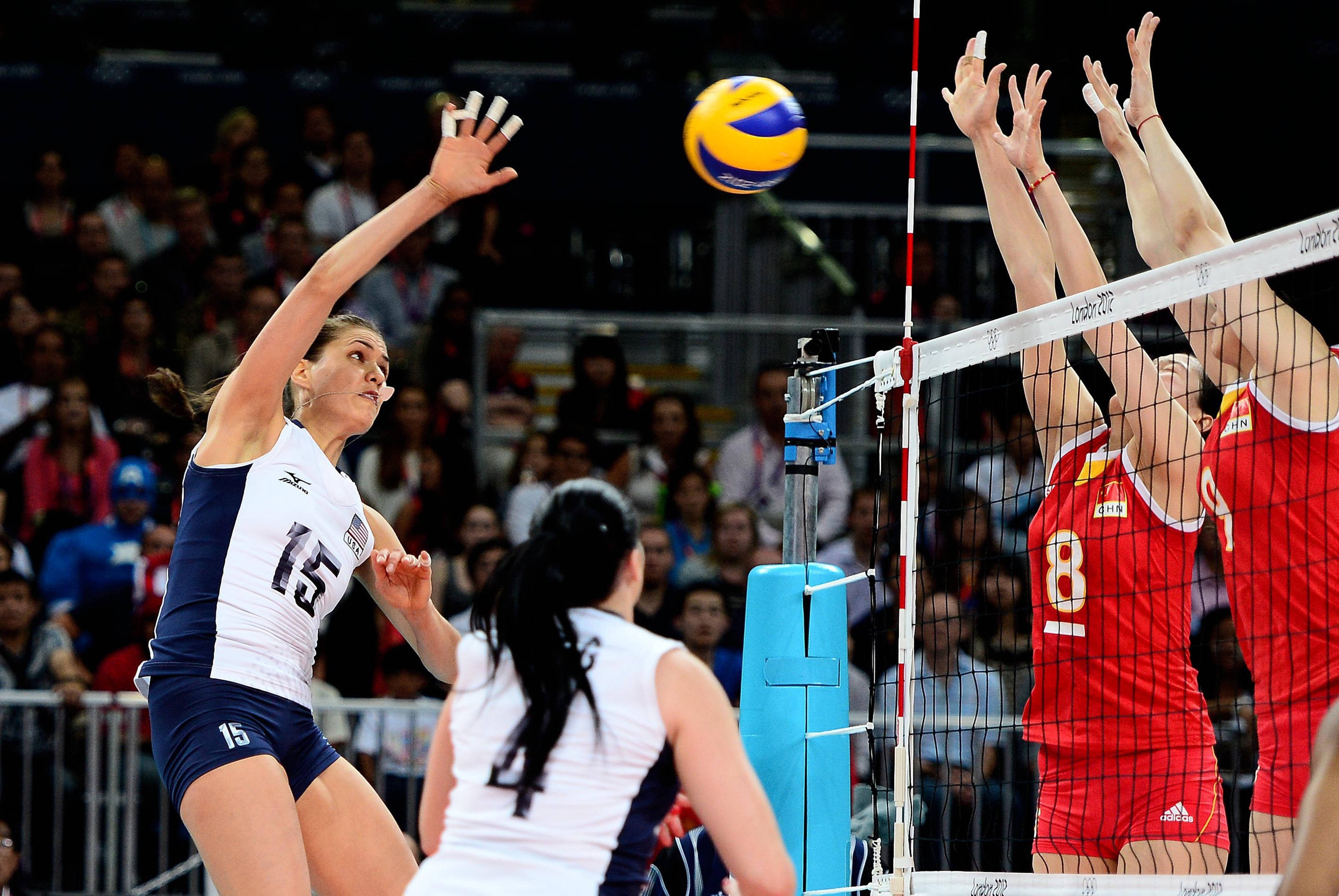 Volleyball 桌布 壁紙 — 一名身穿白色和藍色球衣的女排球員將球扣過網，而身穿紅色的對手試圖攔網。