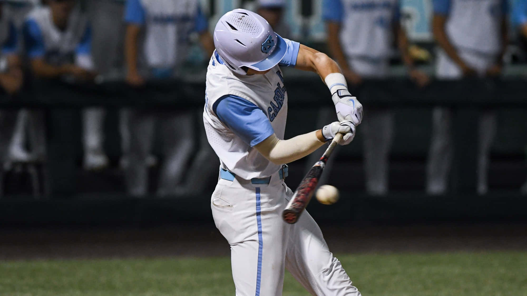 College Baseball 桌布 壁紙 — 一名身穿白色和藍色球衣的棒球選手在球場燈光下揮棒擊球，背景中的隊友模糊不清。
