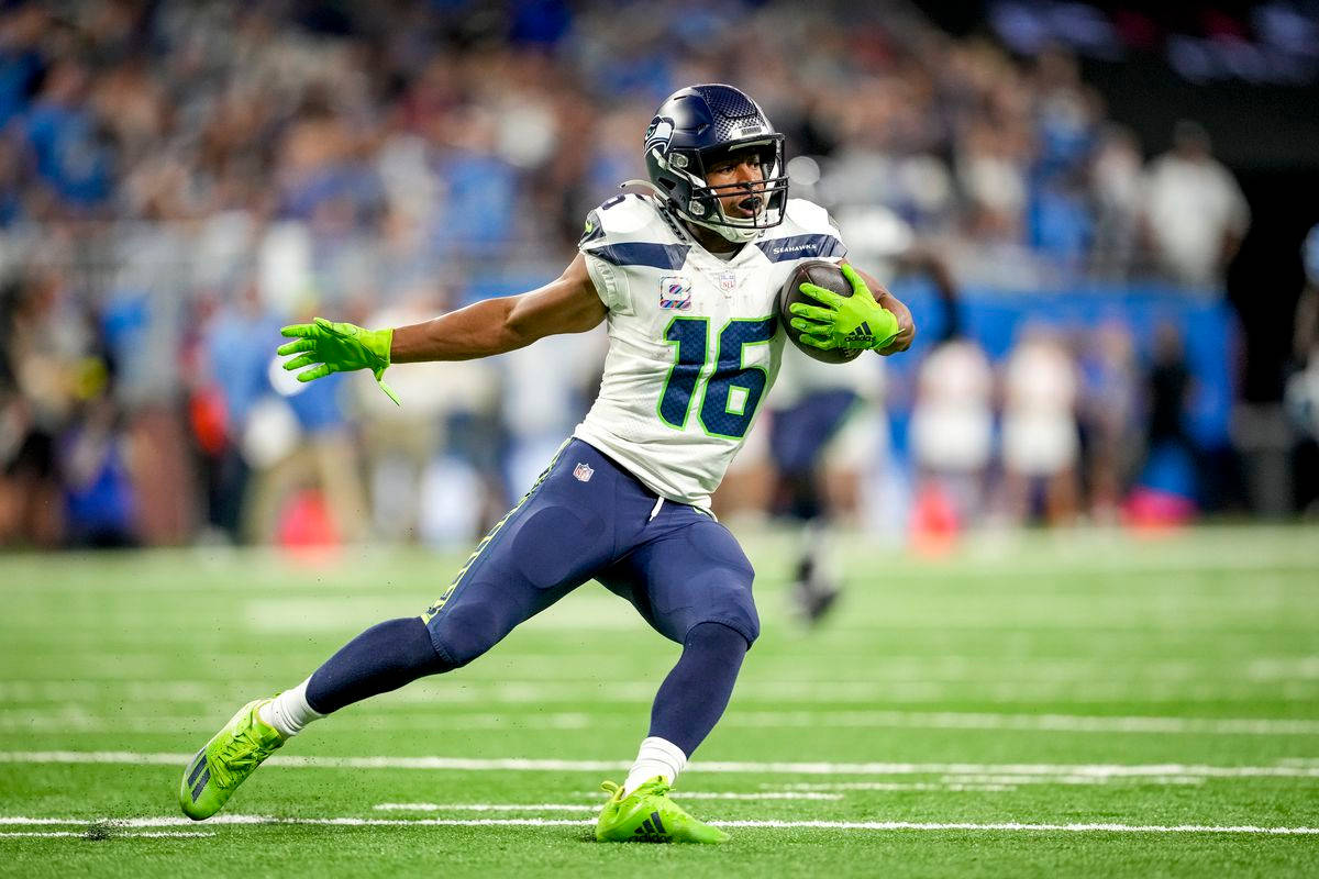Tyler Lockett 桌布 壁紙 — 一名身穿白色和藍色球衣的西雅圖海鷹隊美式足球員在夜間比賽中，在球場燈光下帶球衝刺。