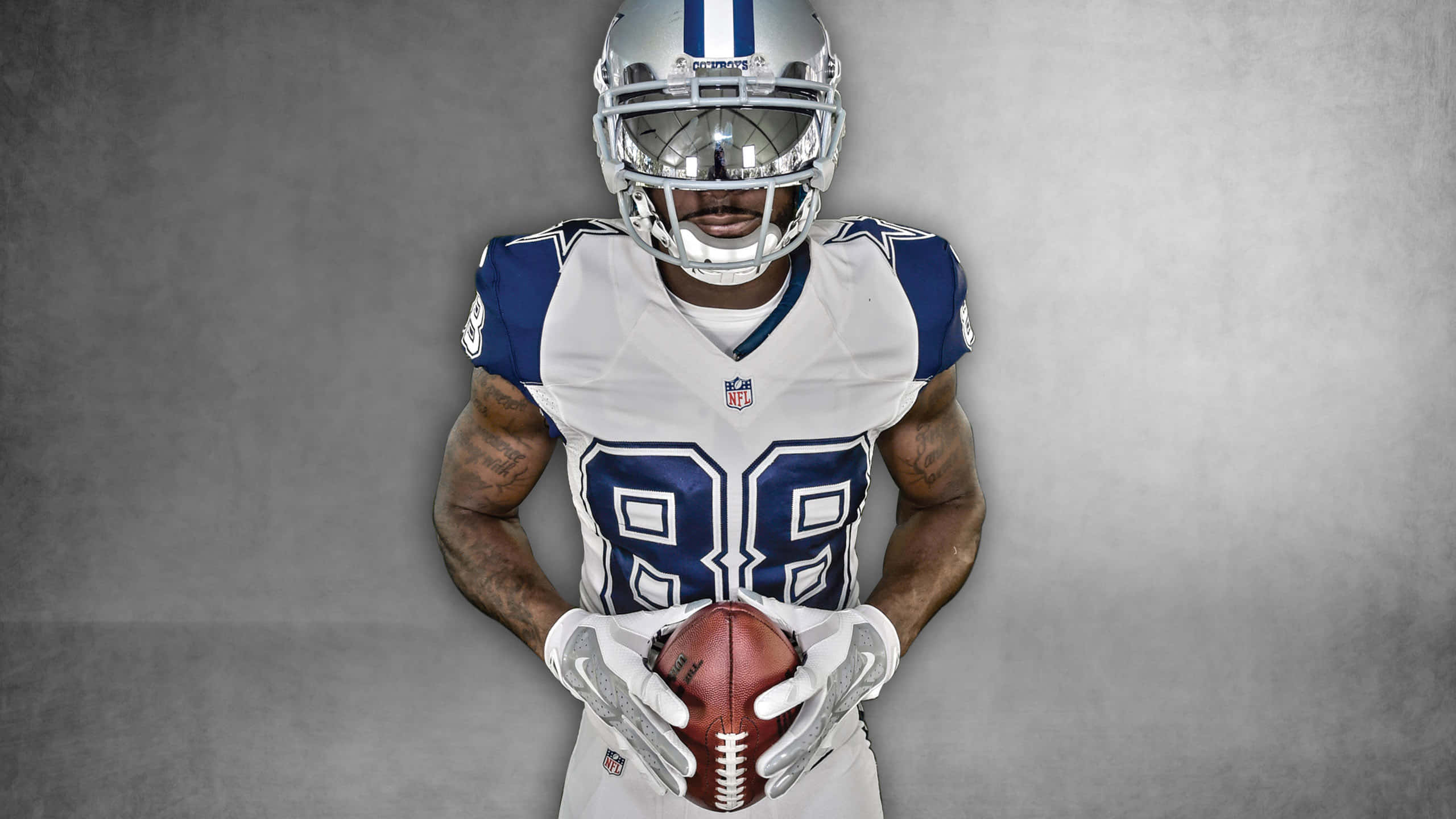 Dallas Cowboys Players 桌布 壁紙 — 一名身穿白色和藍色球衣的達拉斯牛仔隊美式足球員，在灰色攝影棚背景下抱著一個美式足球。