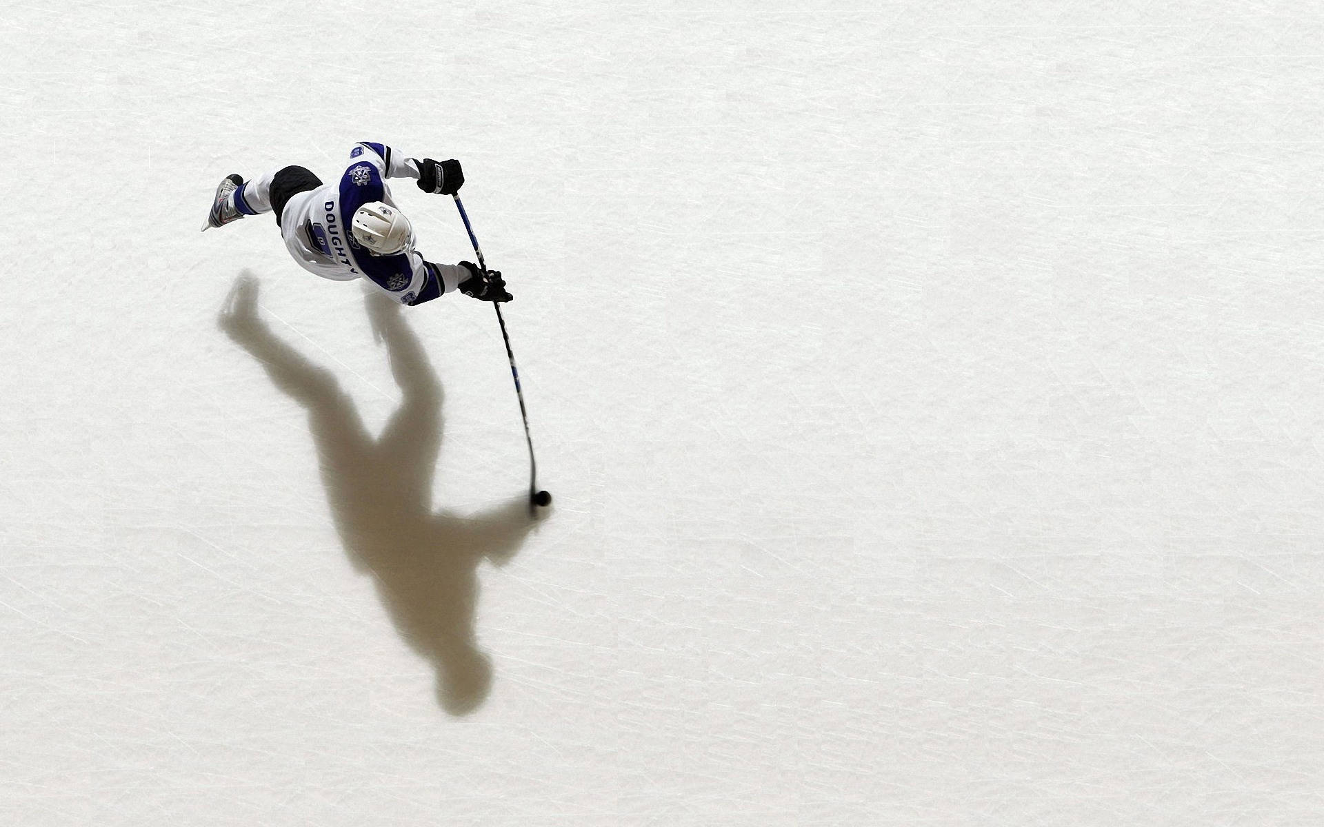 hockey 桌布 壁紙 — 一名身穿白色和藍色裝備的曲棍球運動員在冰上滑行，在冰場上投下長長的影子。