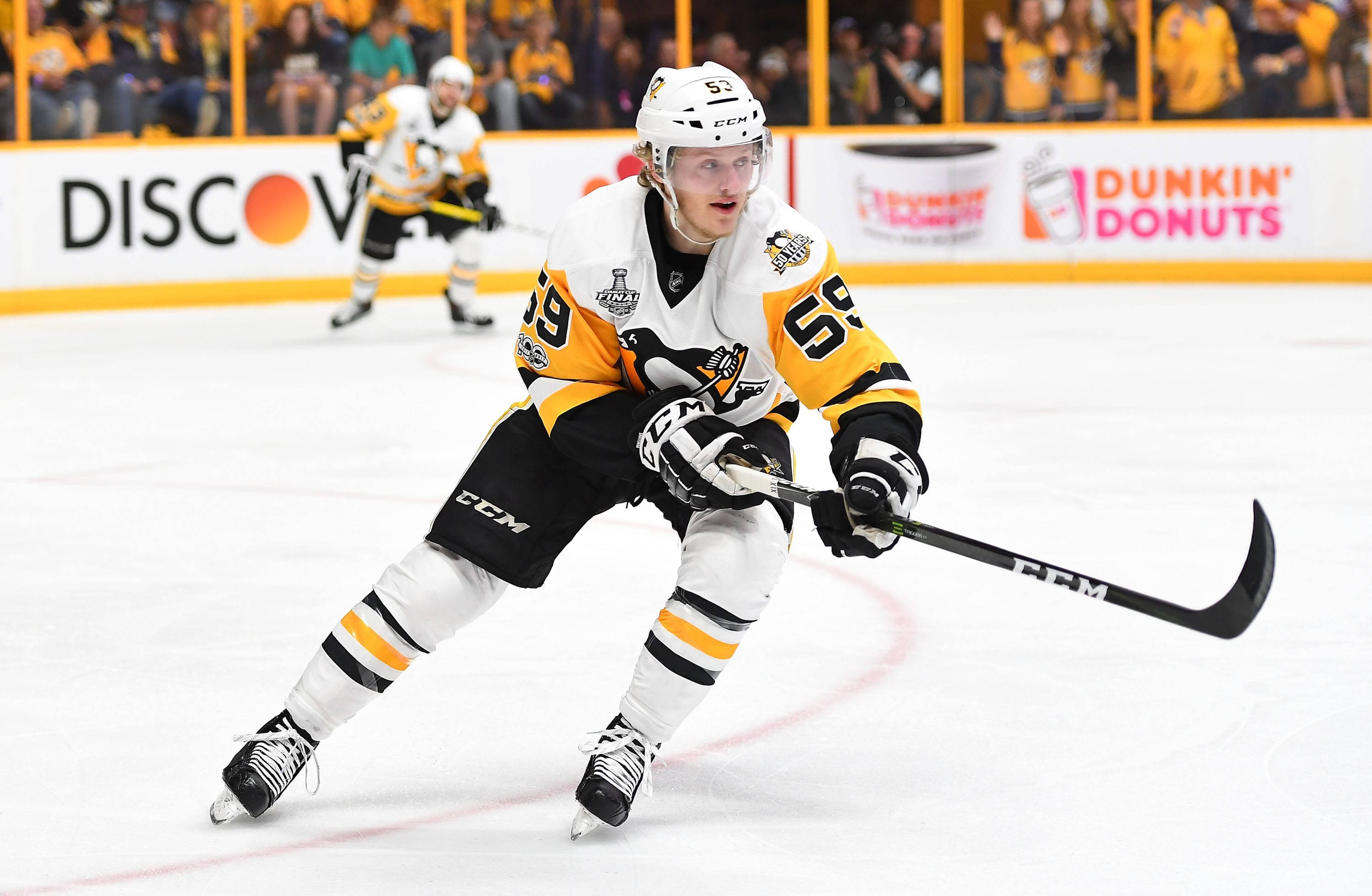 hockey 桌布 壁紙 — 一名身穿白色和金色球衣的冰球運動員在明亮的場館燈光下，在比賽中帶著冰球在冰上激烈滑行。