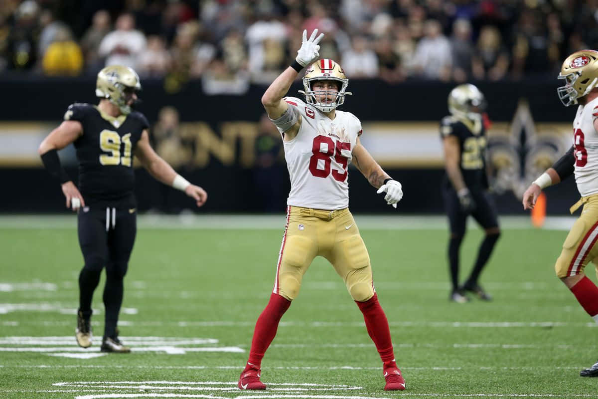 George Kittle 桌布 壁紙 — 一名身穿白色和金色球衣的舊金山四九人美式足球員在綠色球場上舉手慶祝比賽。