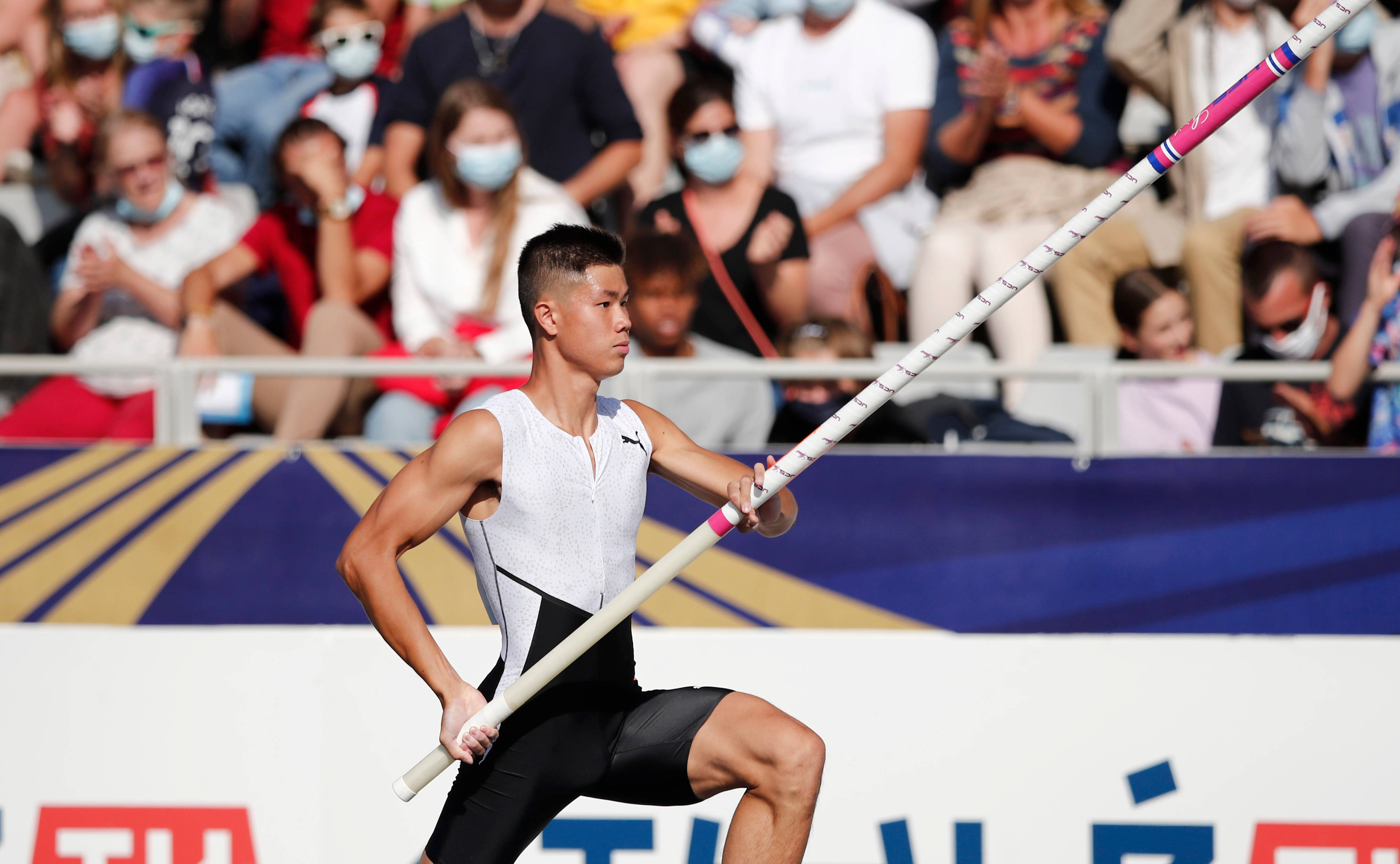 Olympics 桌布 壁紙 — 一名身穿白色和黑色制服的男性撐竿跳運動員，在體育館內專注衝刺，手持粉紅色和白色的竿，背景有戴著口罩的觀眾。