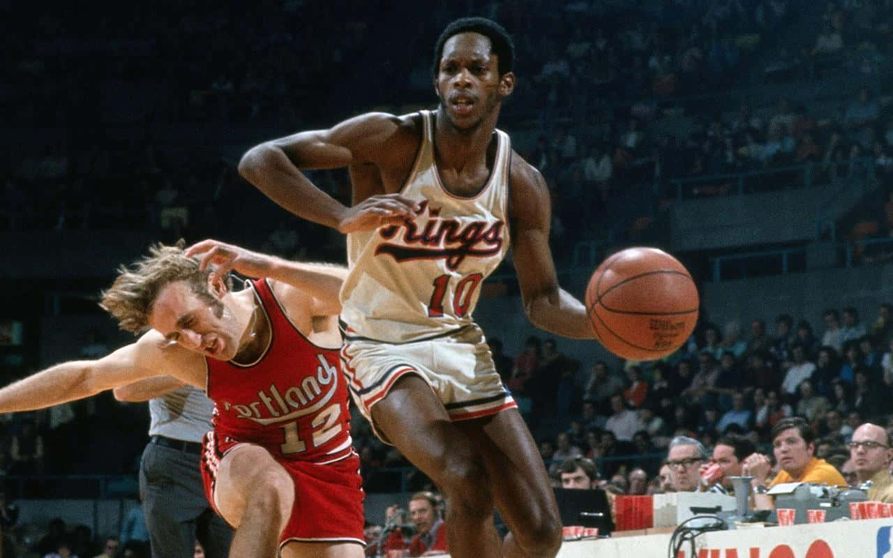 NBA 桌布 壁紙 — 一名身穿白色國王隊球衣的籃球運動員在擠滿觀眾的場館中運球越過紅色波特蘭拓荒者隊的防守球員。
