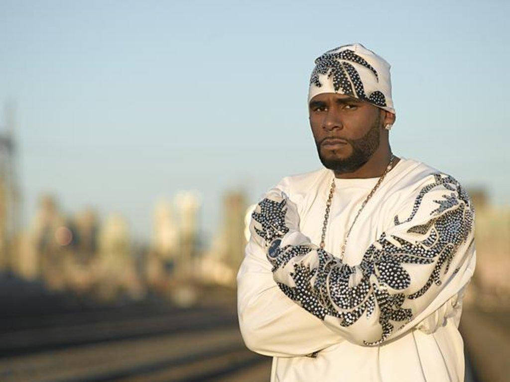 r kelly 桌布 壁紙 — 一名身穿白色圖案服裝和頭巾的男子，在黃金時段以雙臂交叉的姿勢自信地站在模糊的城市天際線前。