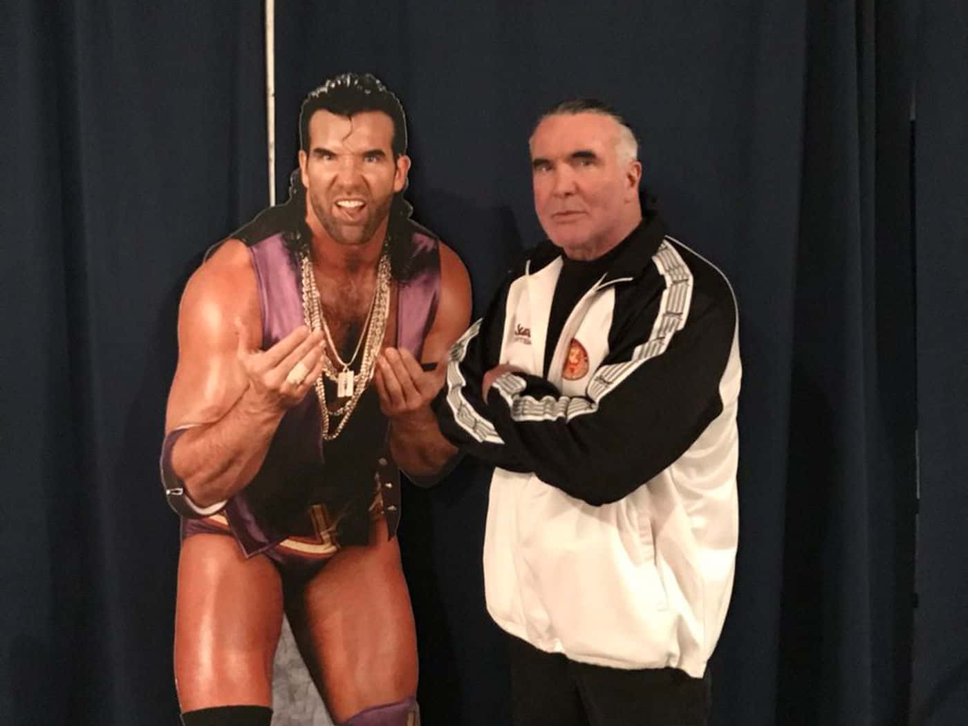 WWE 桌布 壁紙 — 一名身穿白色外套的男子站在一個真人大小的肌肉摔角手剪紙旁邊，該剪紙穿著紫色裝備，背景是深色窗簾。