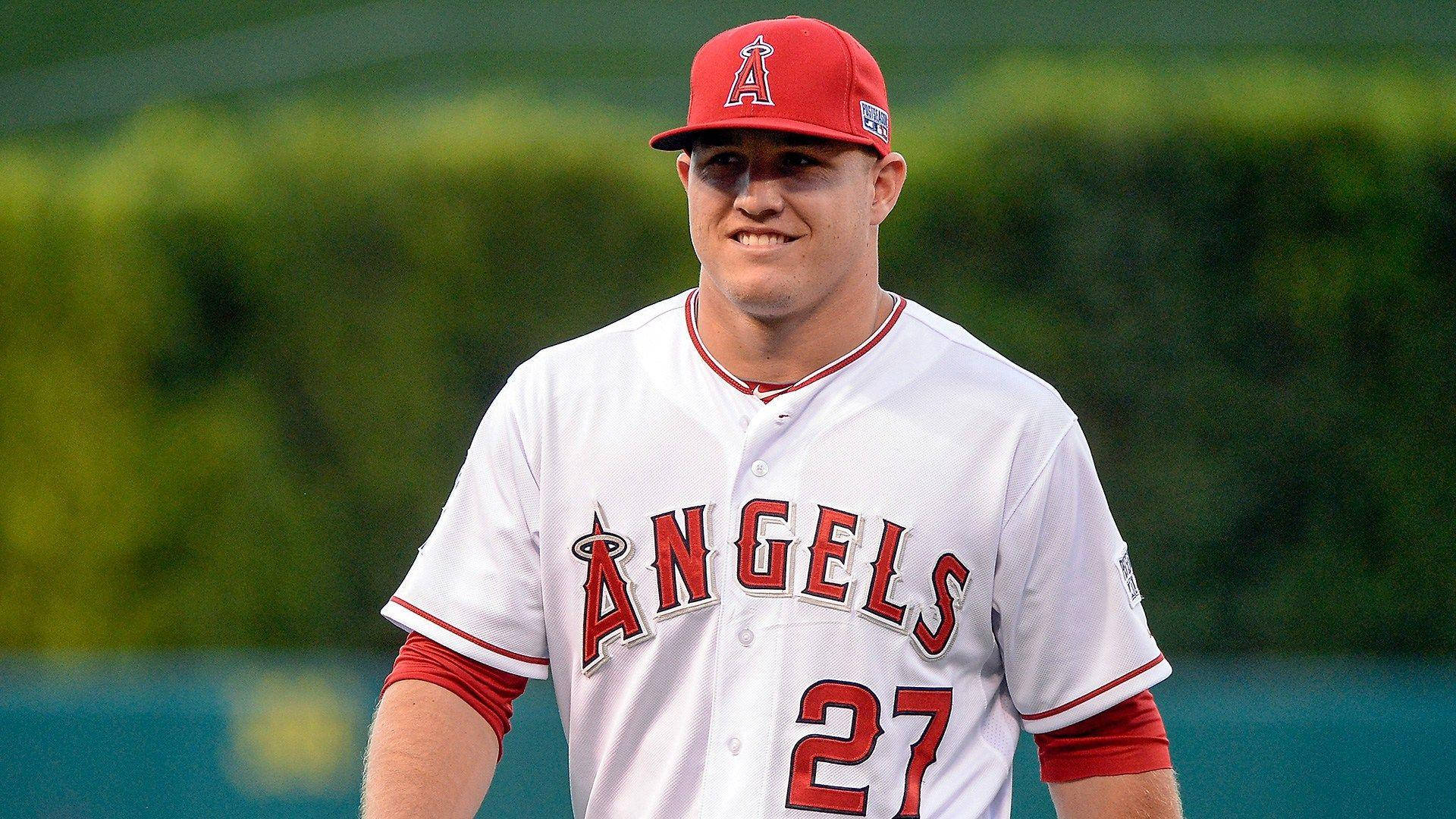 Mike Trout 桌布 壁紙 — 一名身穿白色天使隊球衣、紅色27號的棒球選手在陽光普照的球場上自信地微笑。