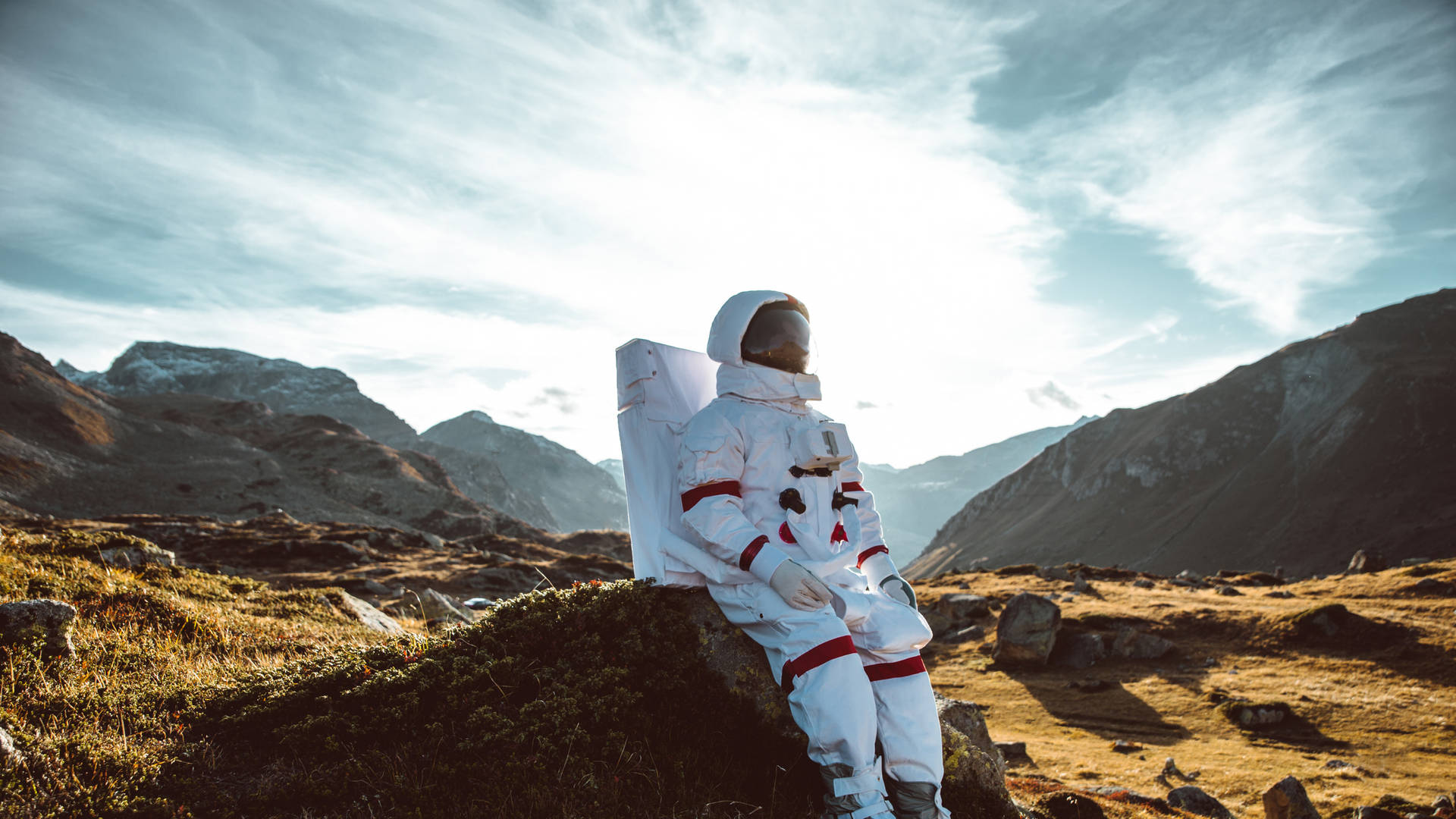 壁紙 — 一名身穿白色太空衣的太空人獨自坐在明亮的天空下，崎嶇山脈中一塊長滿苔蘚的岩石上。
