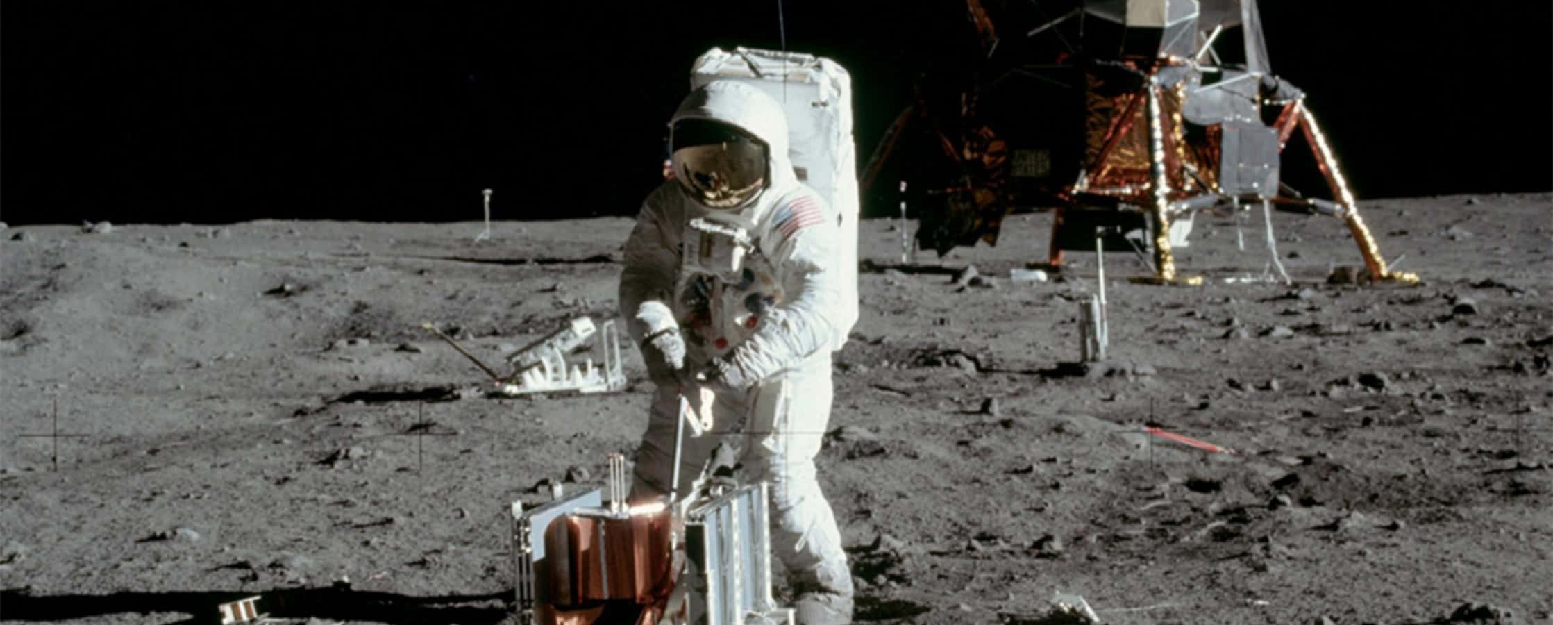Moon Landing 桌布 壁紙 — 一名身穿白色太空衣的太空人站在月球灰色的表面上，旁邊是月球登陸艙，背景是黑色的天空。